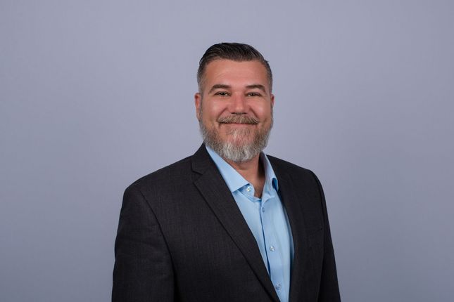Man with a short, spiky haircut and stubble, smiling. Wearing a blazer and light blue shirt, with a blue background.