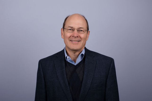 Man with glasses smiles, wearing a burgundy sweater over a white collared shirt, in front of a blue background.