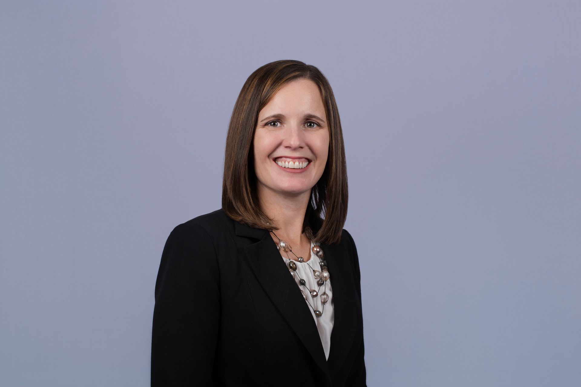 Woman with brown hair, wearing a black blazer and smiling, against a blue background.