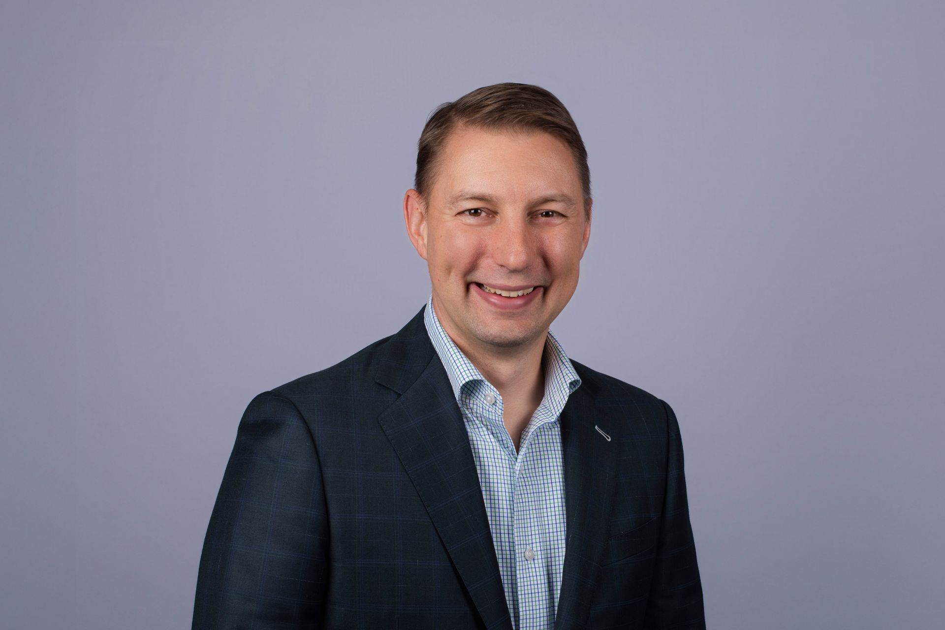 Man in a blue suit smiles, looking directly at the camera against a light blue background.