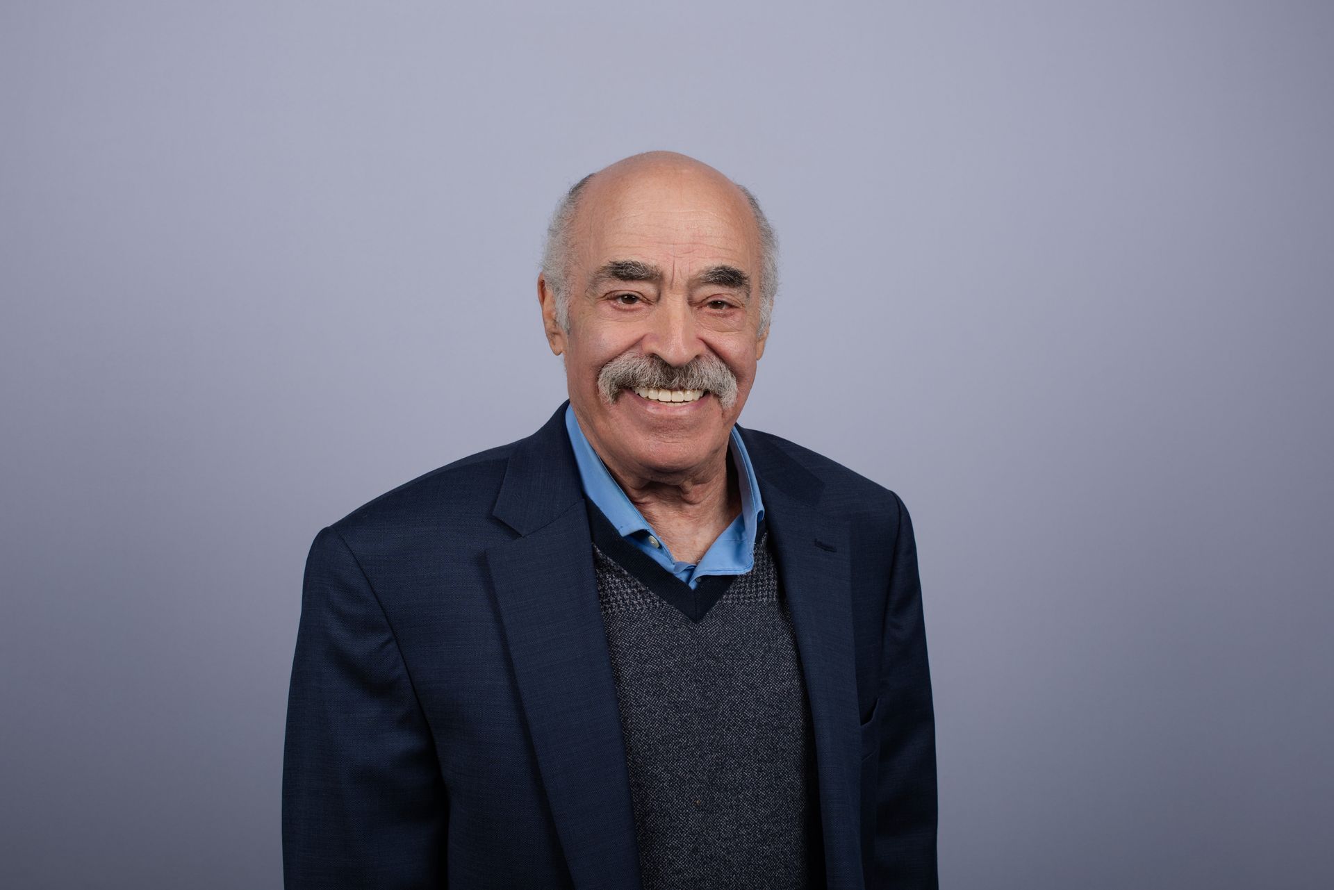 Man with a short, spiky haircut and stubble, smiling. Wearing a blazer and light blue shirt, with a blue background.