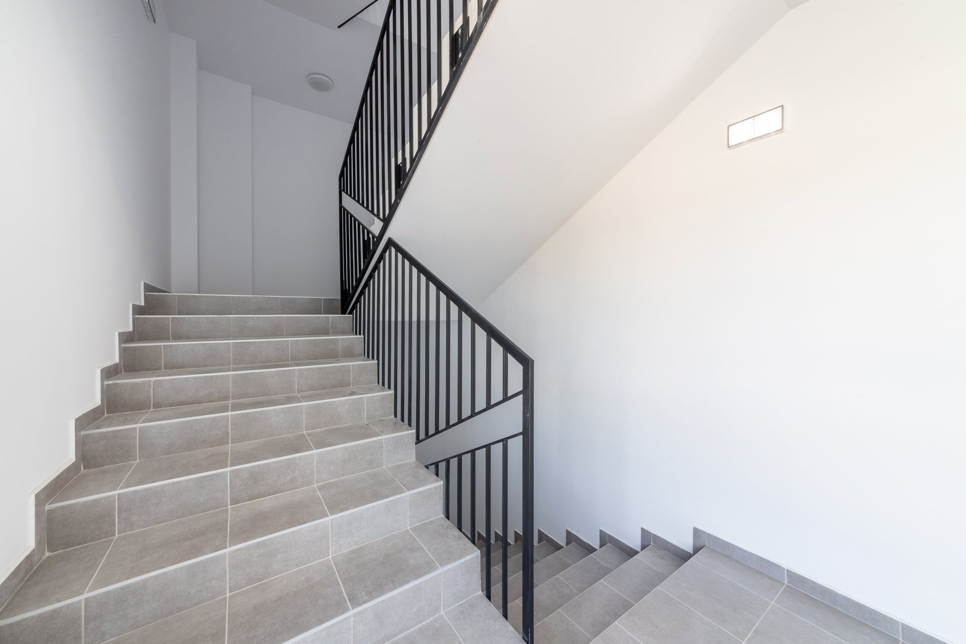 Gray tiled stairs with black metal handrails leading upwards, flanked by white walls.