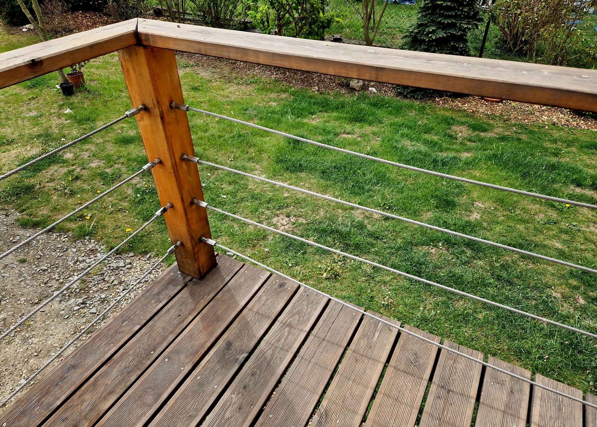 Wooden deck railing with horizontal steel cables, in a yard with grass.