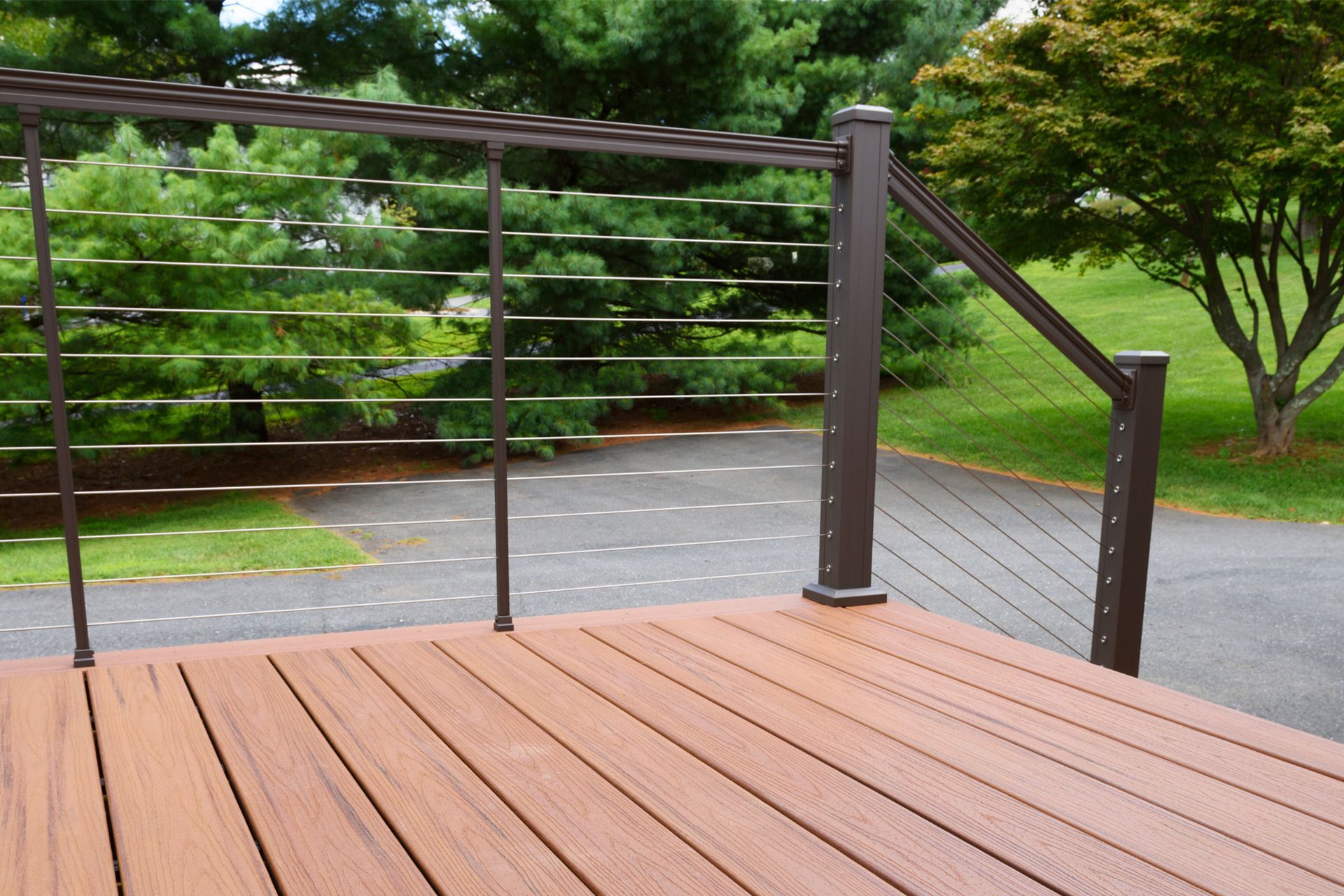 Brown deck with cable railing and overhead structure, overlooking a paved area and green lawn.