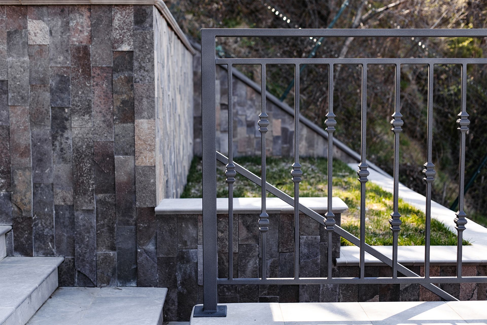 Stone steps with a dark metal railing against a textured stone wall, outdoors.