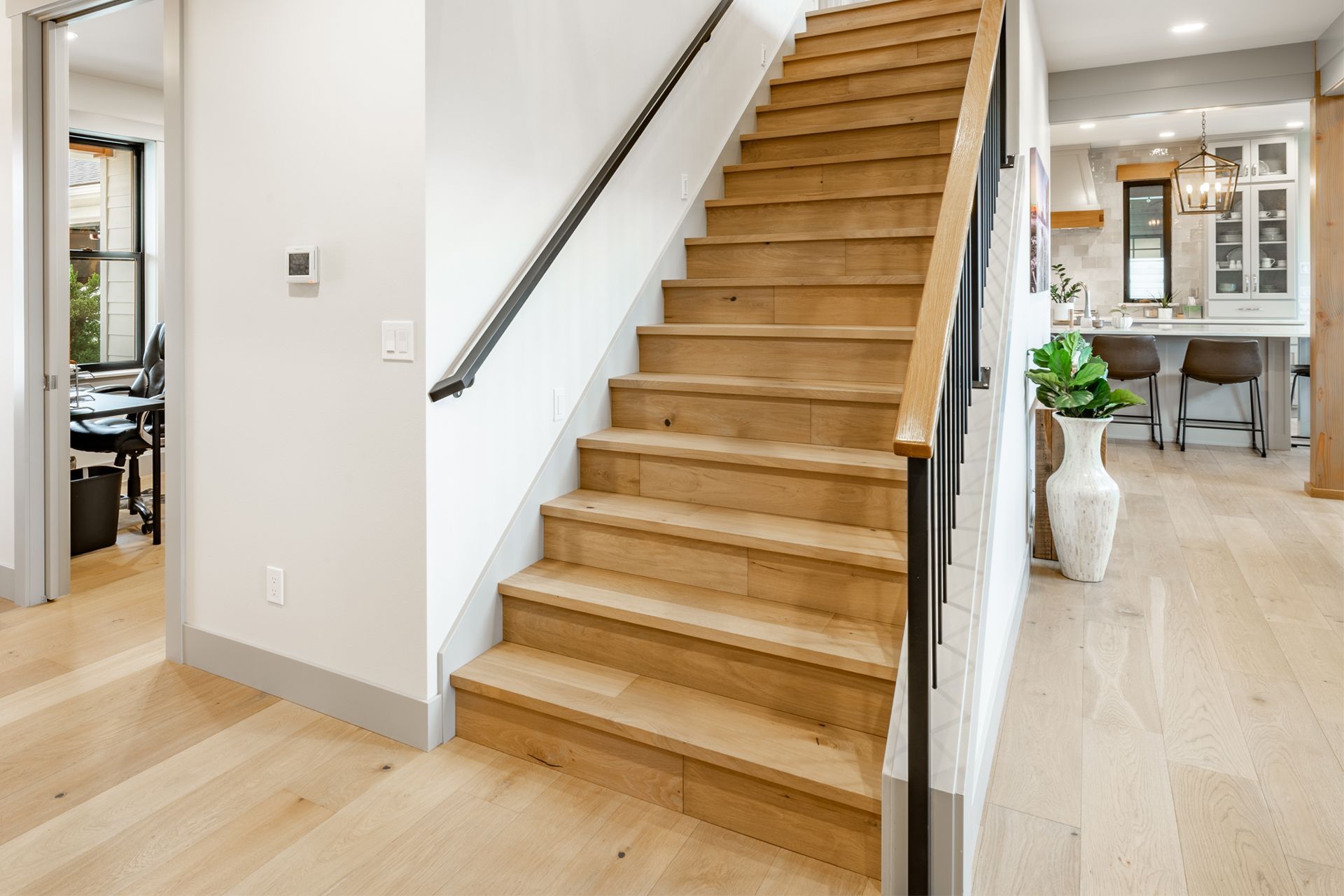 Wooden staircase with black railing and light wood flooring, leading up. Adjacent to an open-concept kitchen.