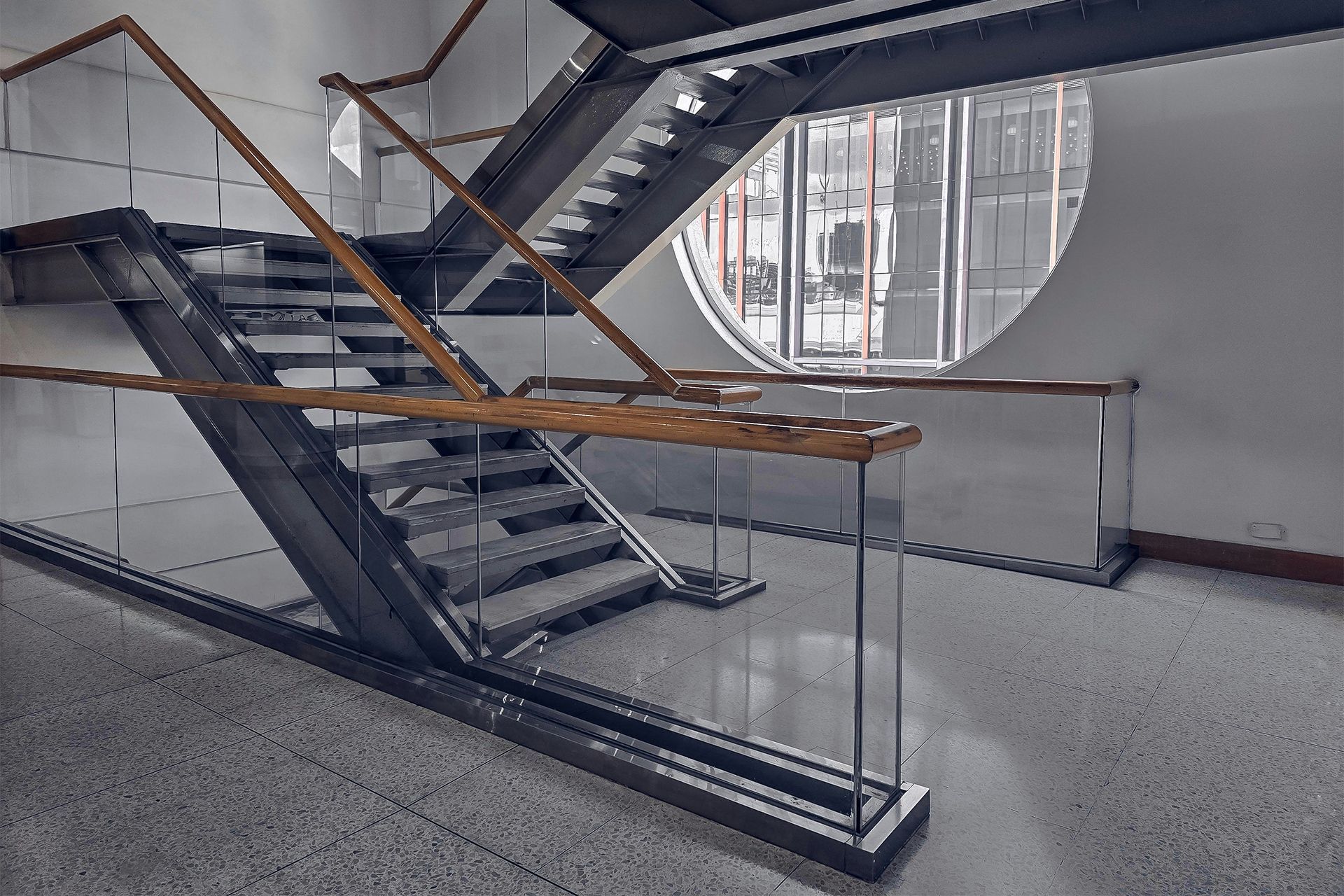 Metal and wood staircase with glass railings, a round window in the background.