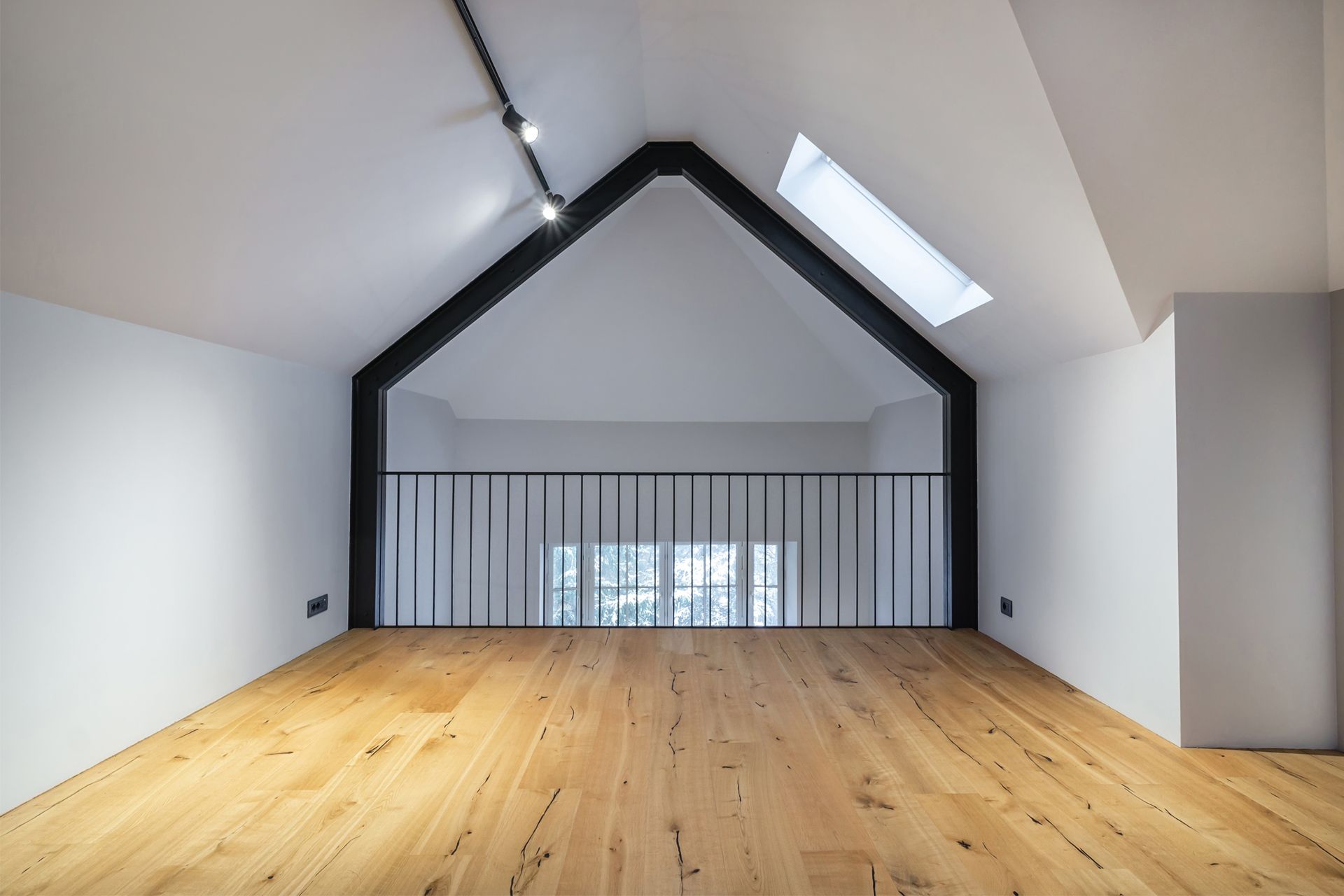 Loft with wooden floor, black railing, skylight, and black structural beam. White walls and ceiling.