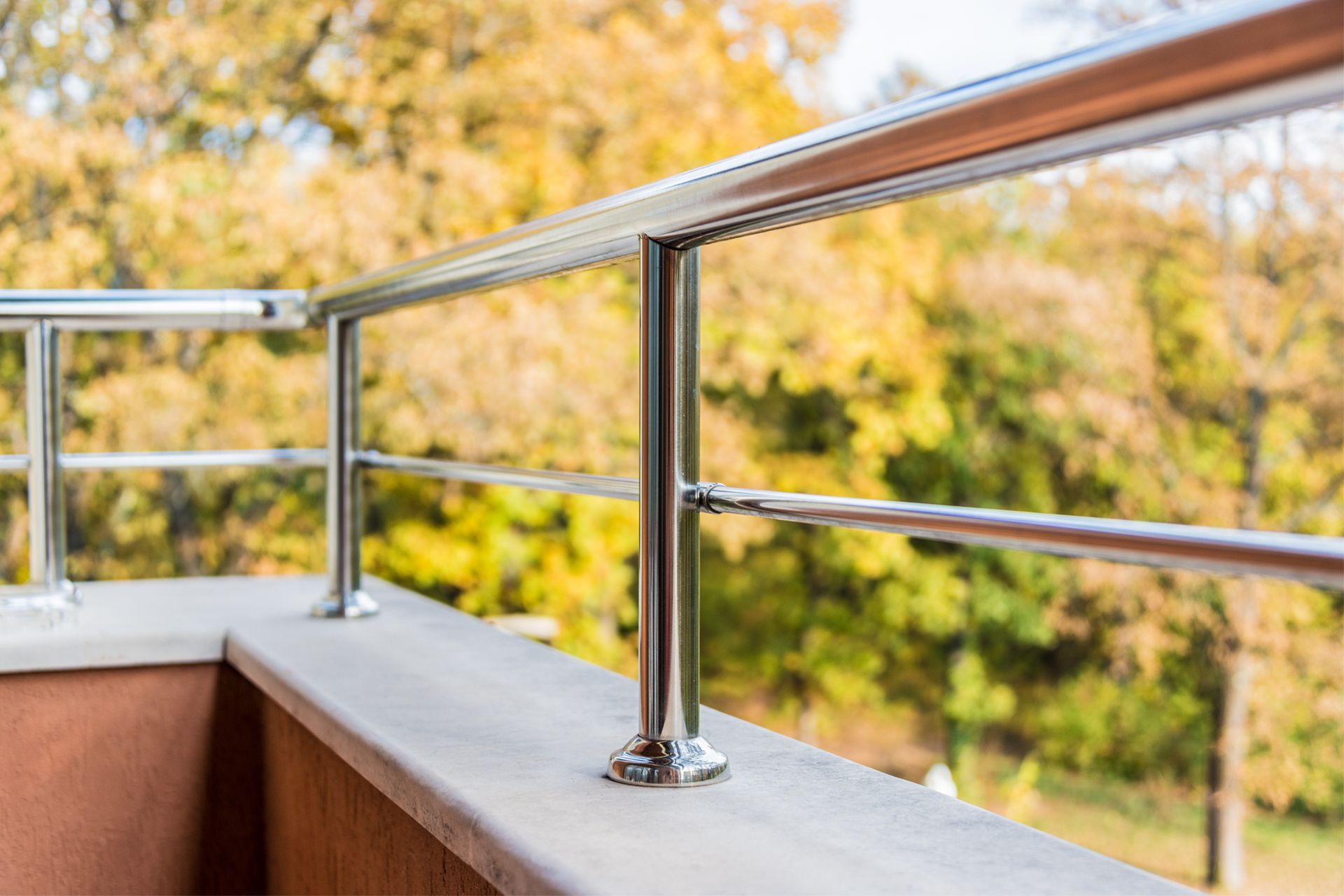 Shiny metal railing on a concrete balcony overlooking blurred autumn trees.