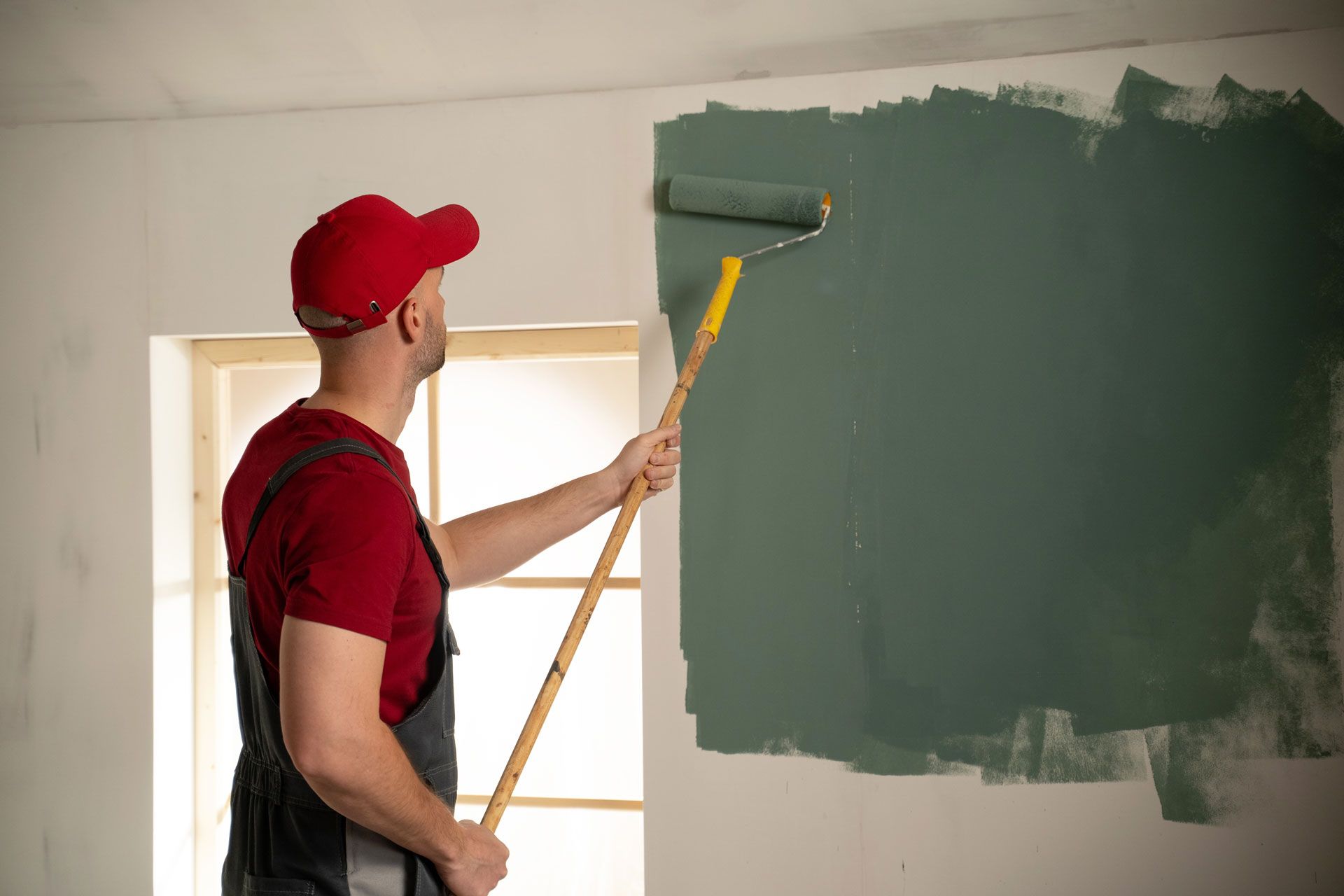 Man in red cap painting a wall green with a roller near a window