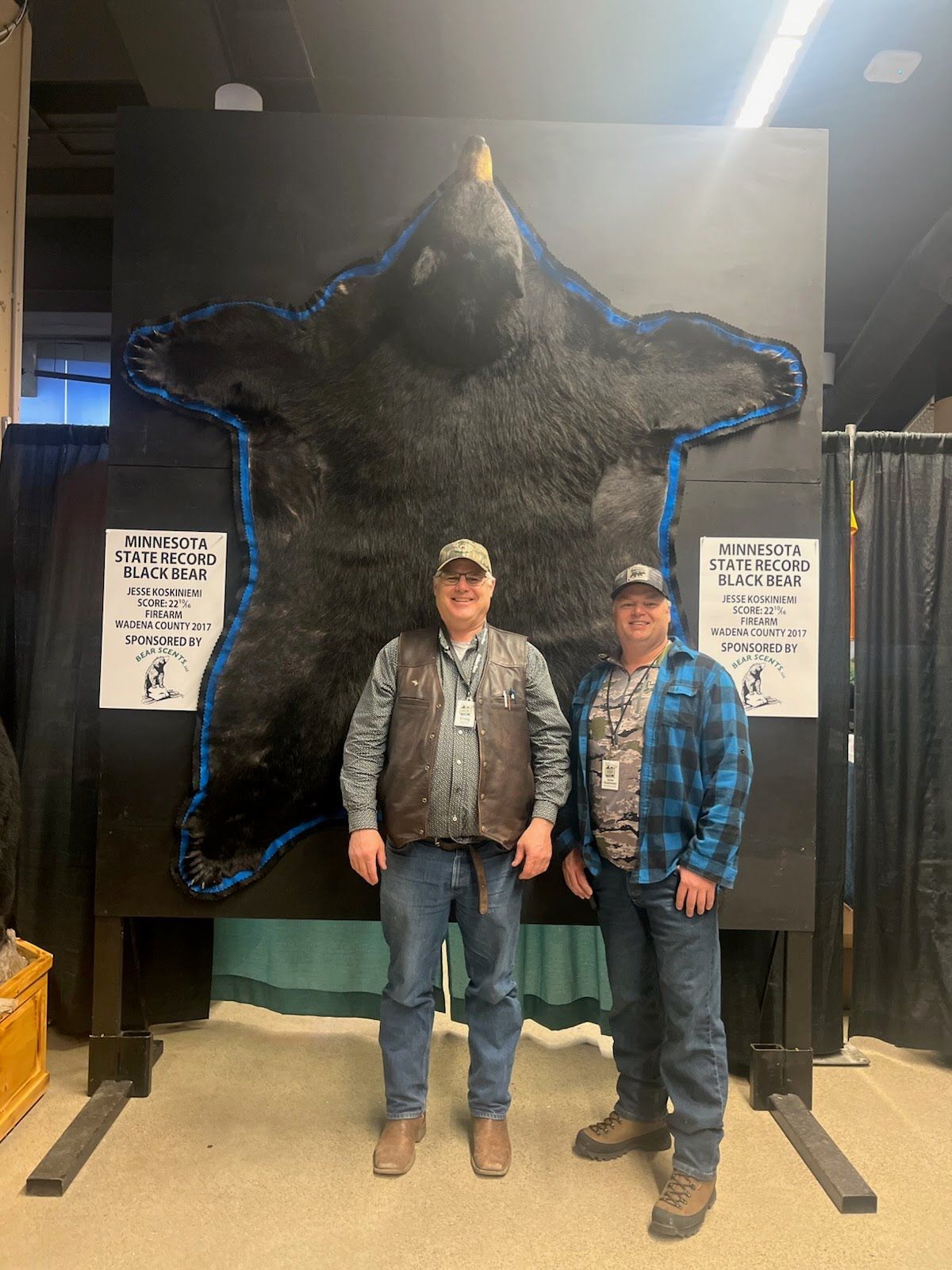 Two men are standing in front of a large black bear rug.