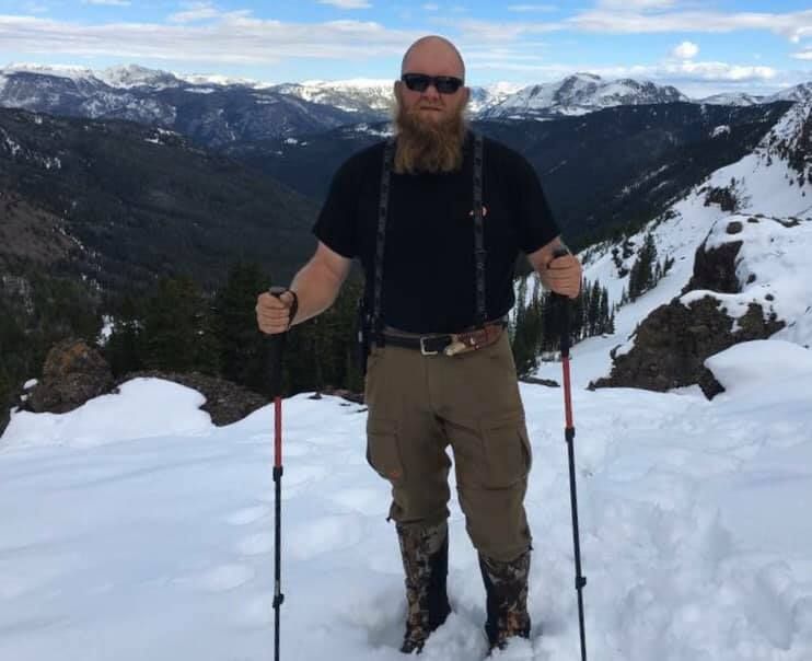 A man with a beard is standing in the snow with hiking poles.