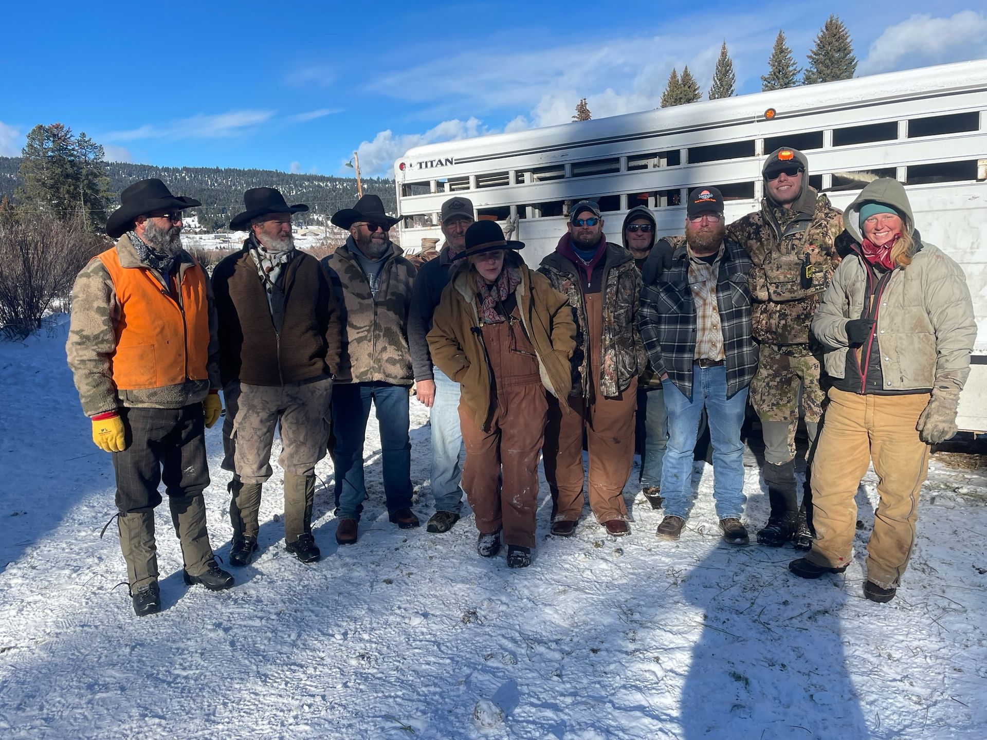 A group of men are standing in the snow in front of a horse trailer.