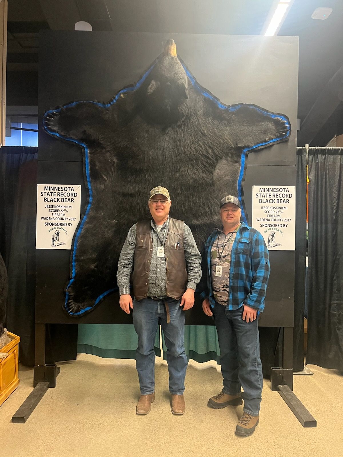 Two men are standing in front of a large black bear rug.
