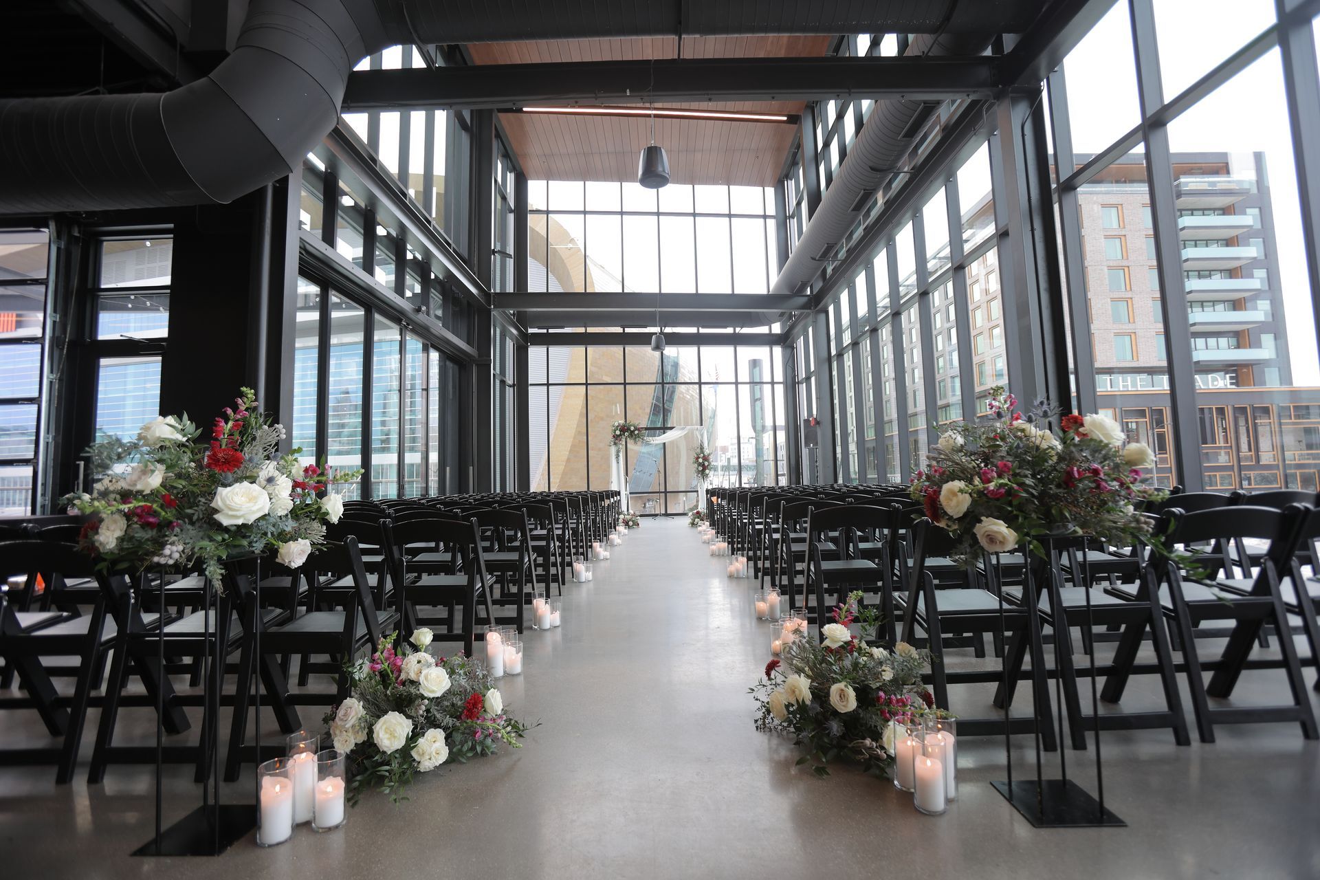 Indoor wedding ceremony setup; black chairs, floral arrangements, candles, floor-to-ceiling windows, city view. With Love, Wedding management, Milwaukee Wedding Coordinator