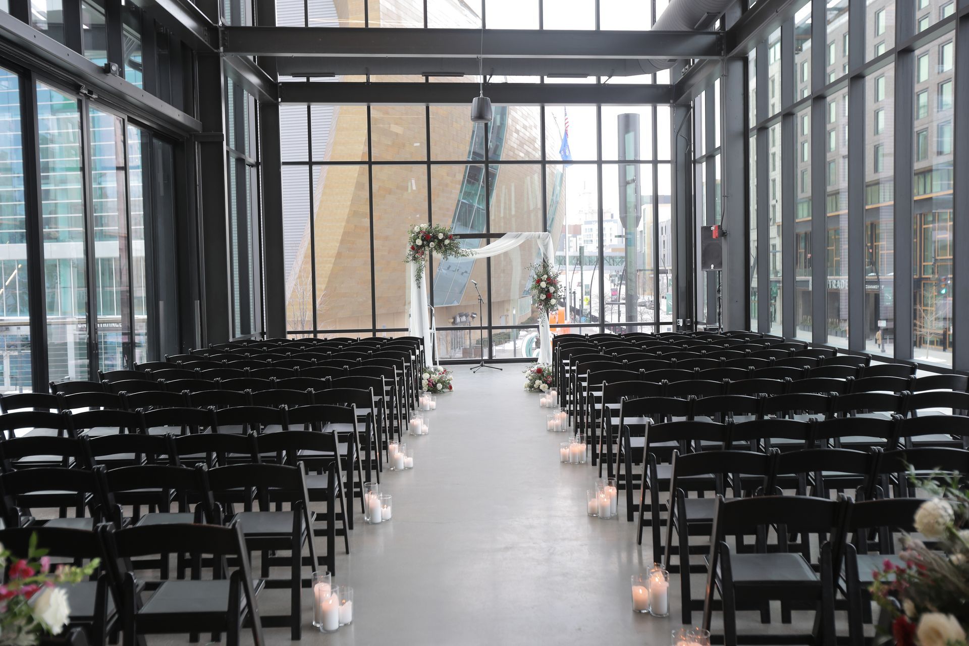 Wedding ceremony setup with black chairs, floral arch, and large windows overlooking city. With Love, Wedding management, Milwaukee Wedding Coordinator