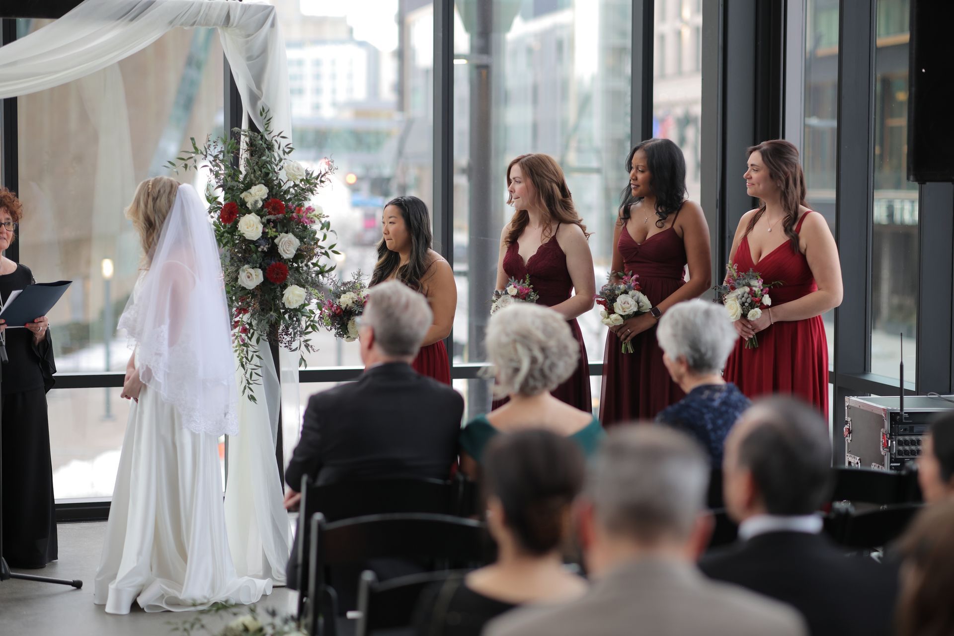 Wedding ceremony in a modern venue. Bride at altar with bridesmaids and guests seated, red floral arrangement, city view.
With Love, Wedding management, Milwaukee Wedding Coordinator
