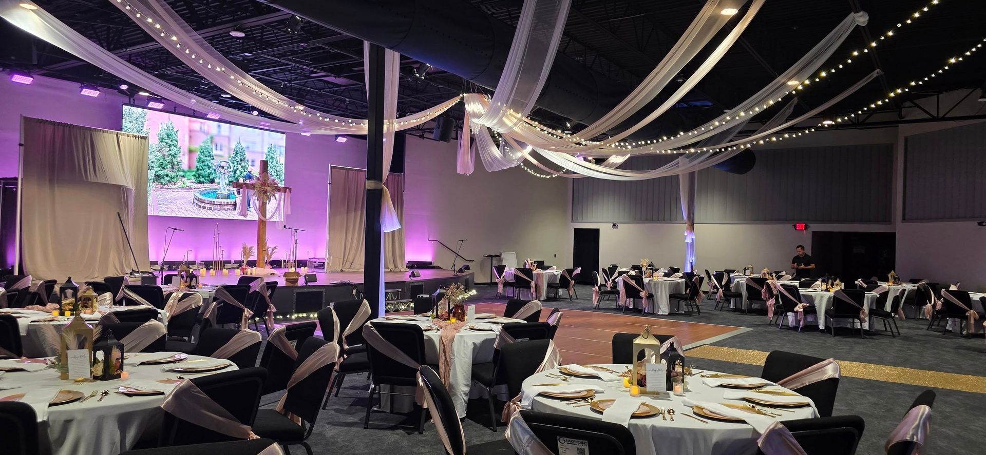 Banquet hall set for an event; tables set, stage with screen and cross, ceiling draped with fabric.