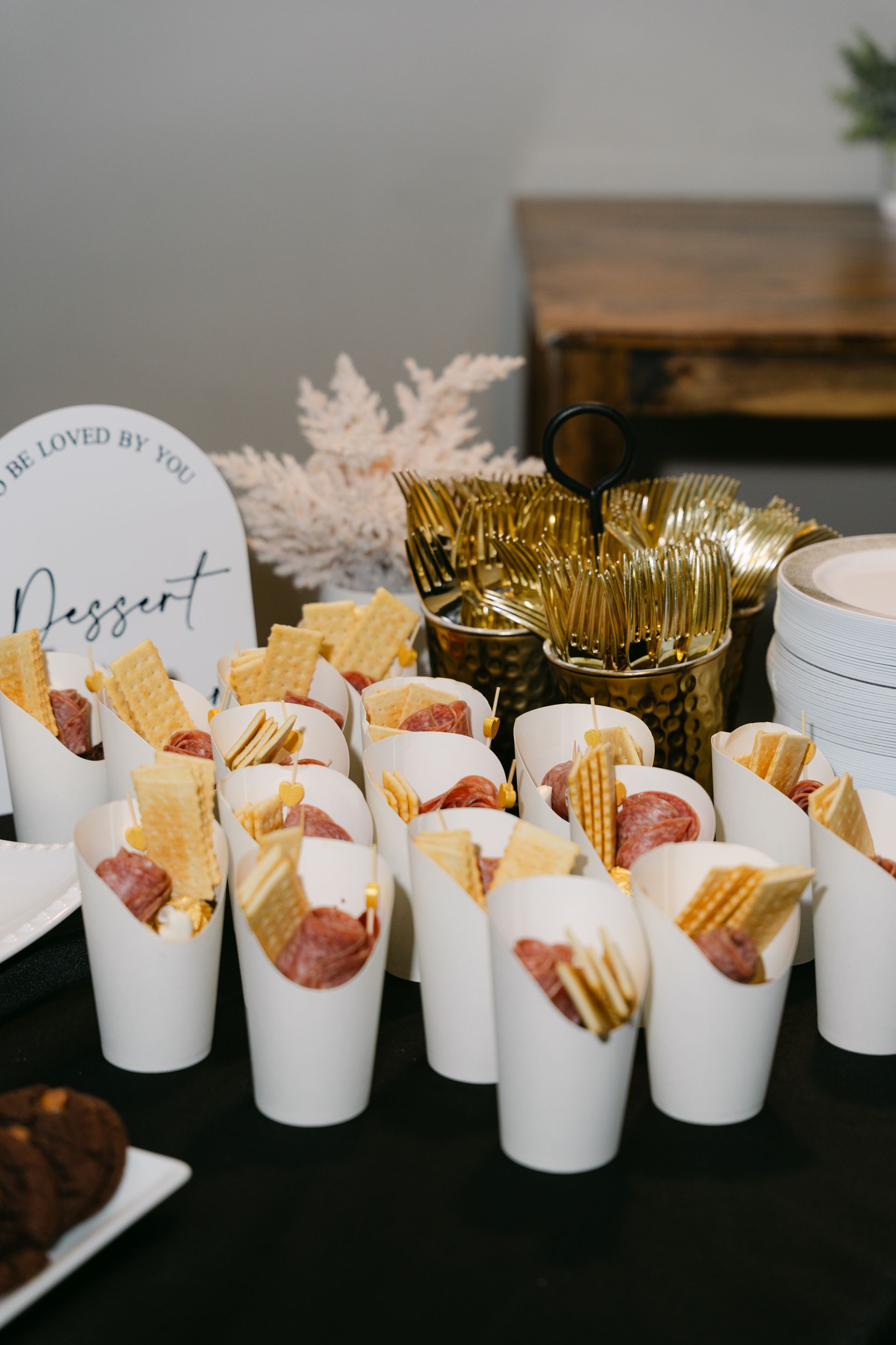 Dessert display: Cups holding sausages and crackers, with gold utensils and sign on a black table.