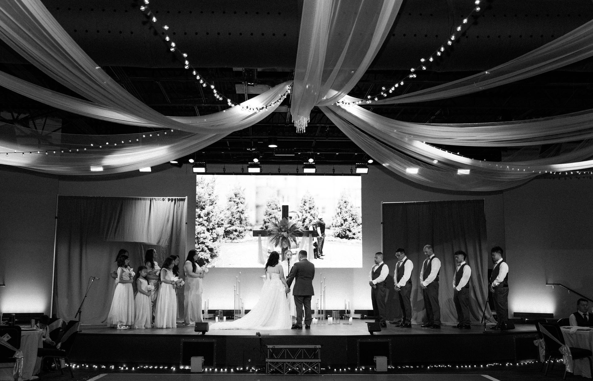 Wedding ceremony on stage with wedding party. Snowy forest backdrop, draping, and string lights.