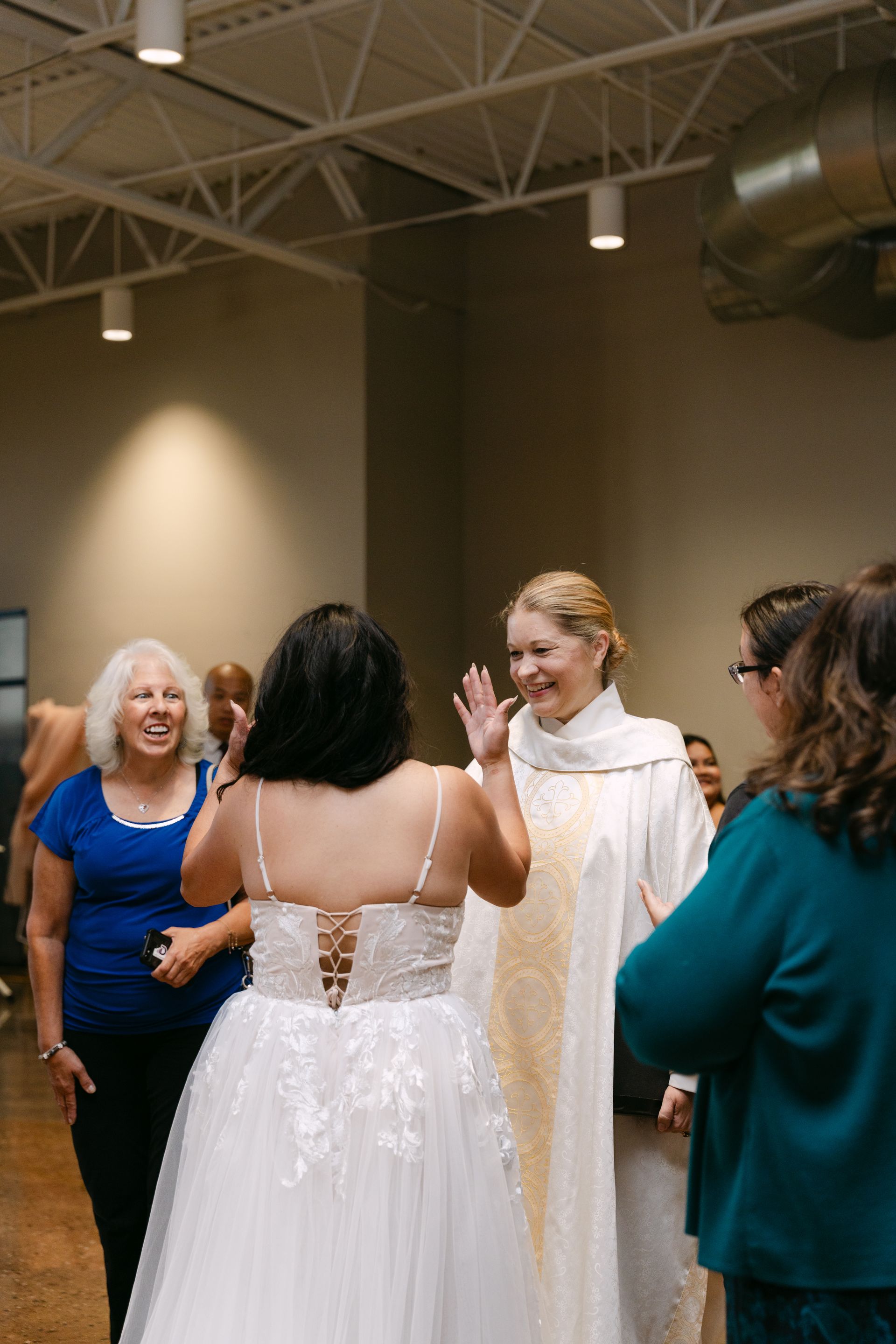 Bride in a white gown and priest in white robe with gold trim greeting guests at an event.