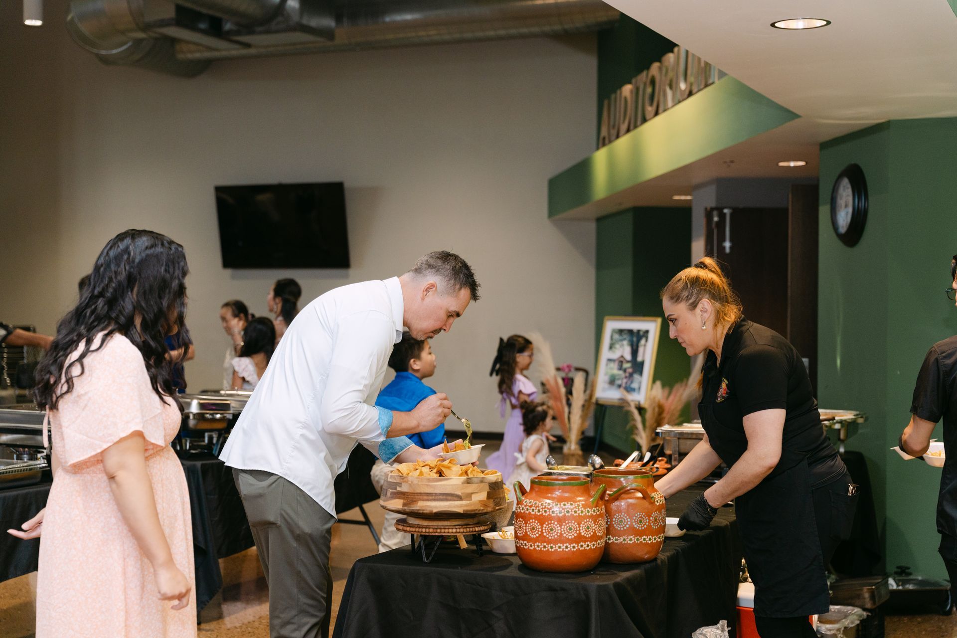 People at a buffet. Man serving food, woman behind the counter, other guests nearby.