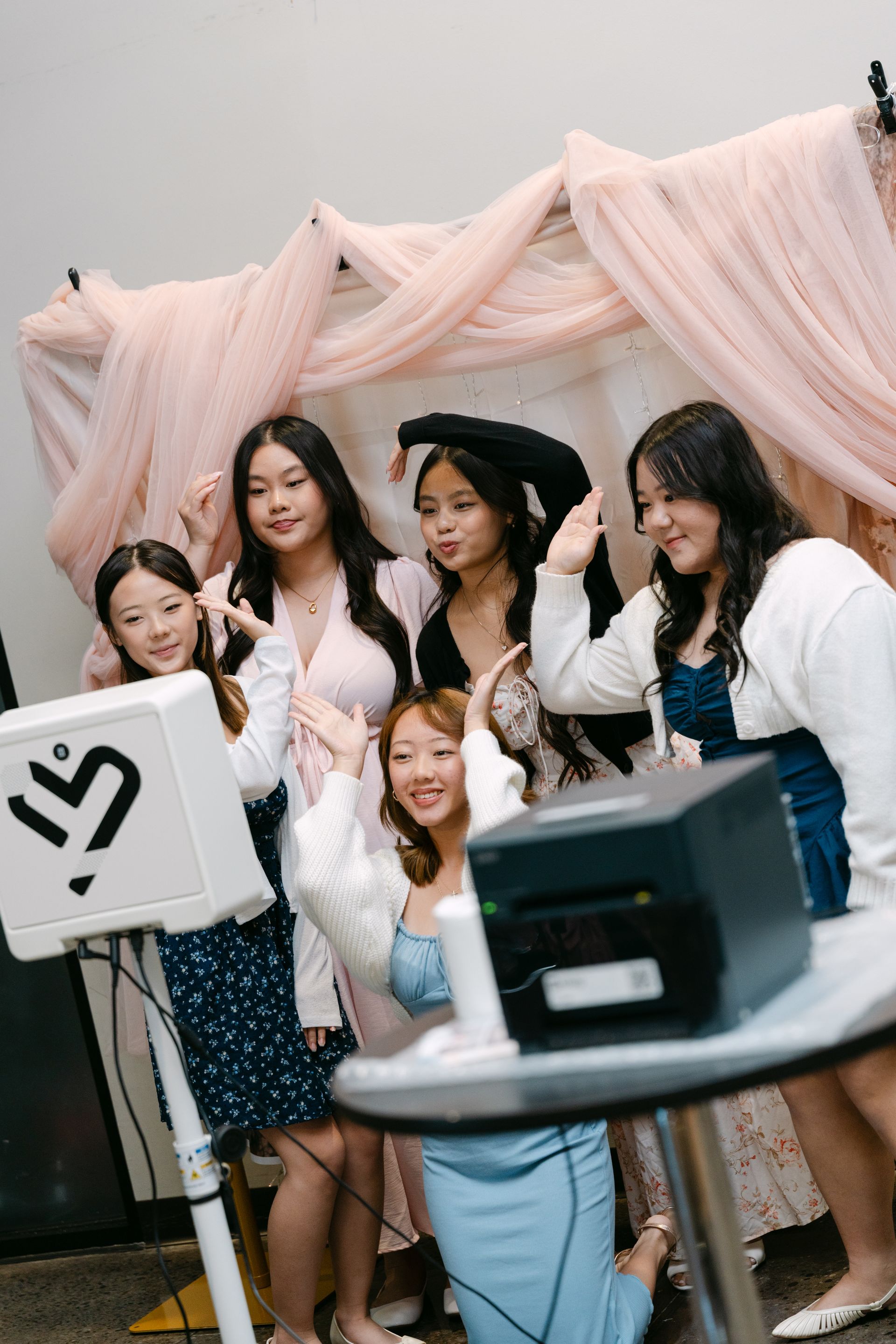 Five young girls pose at a photo booth with pink fabric backdrop. They smile and gesture, some in formal wear.