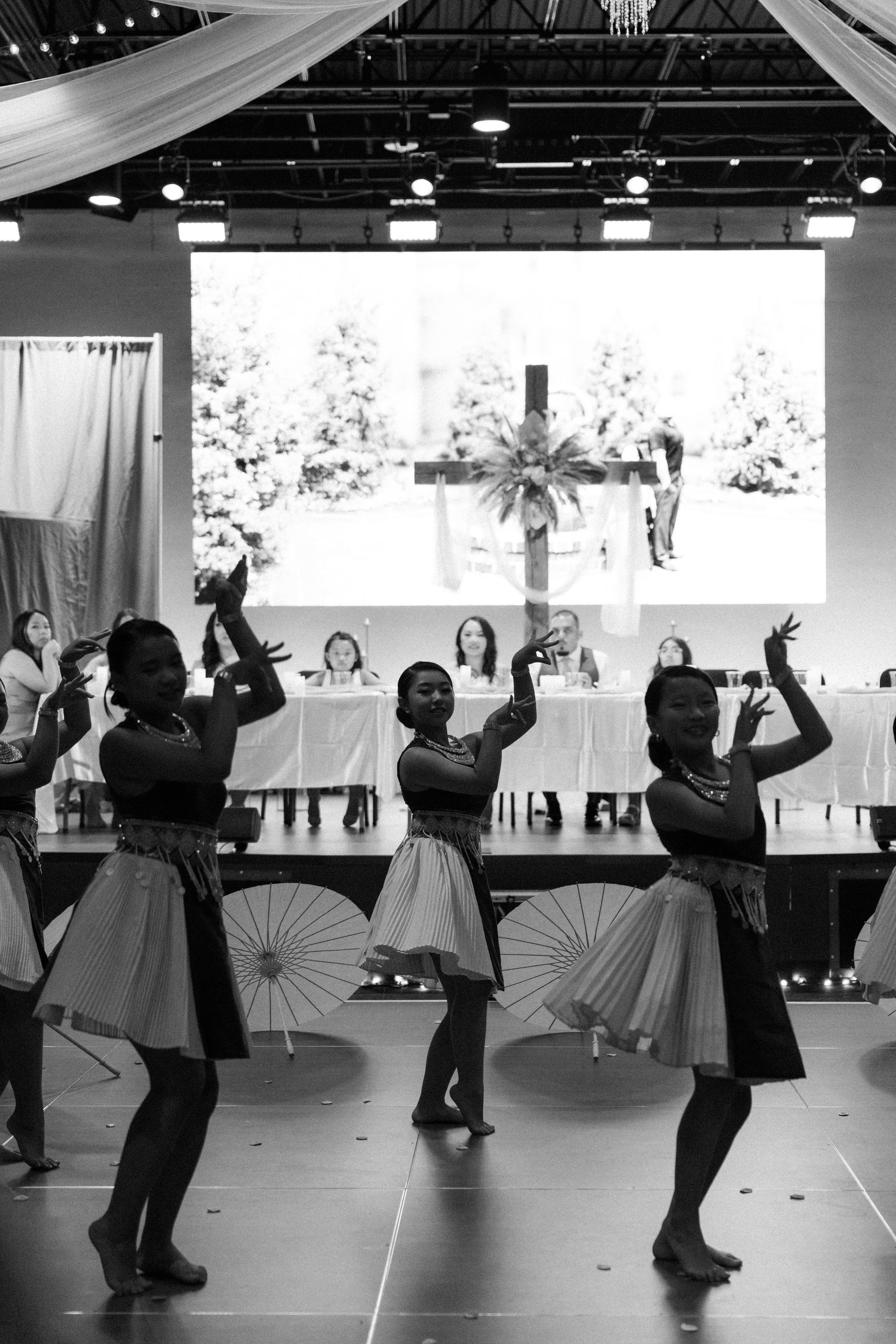 Dancers in skirts perform on stage; backdrop shows a cross. Interior setting.