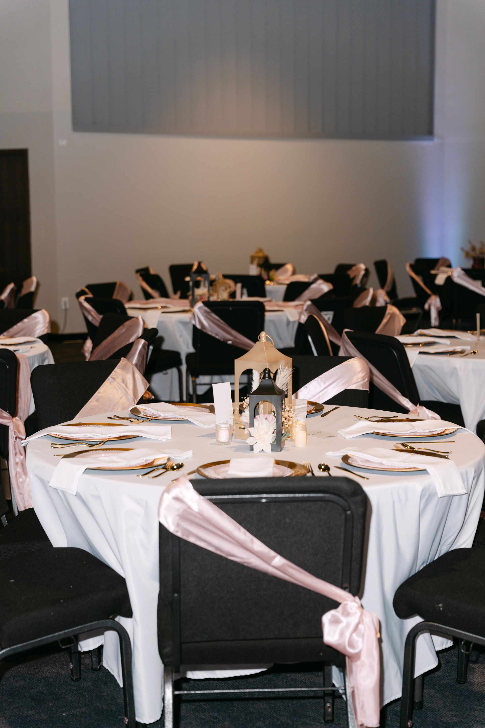Tables set for an event, black chairs with pink sashes, white tablecloths, gold accents, and a candlelit lantern.