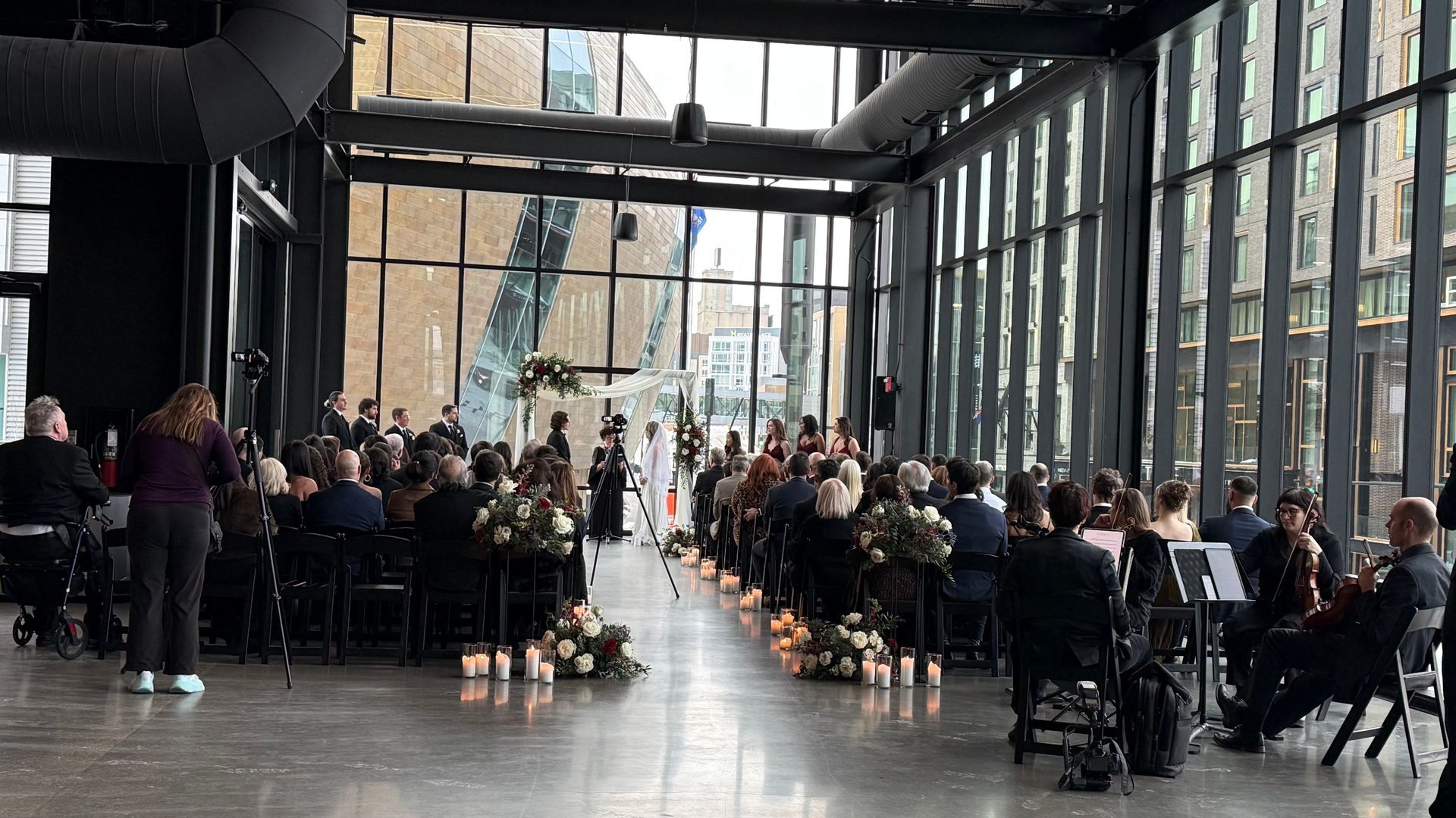 Wedding ceremony in a modern venue. Guests seated, couple at the altar. Large windows, natural light, floral decorations.
With Love, Wedding management, Milwaukee Wedding Coordinator