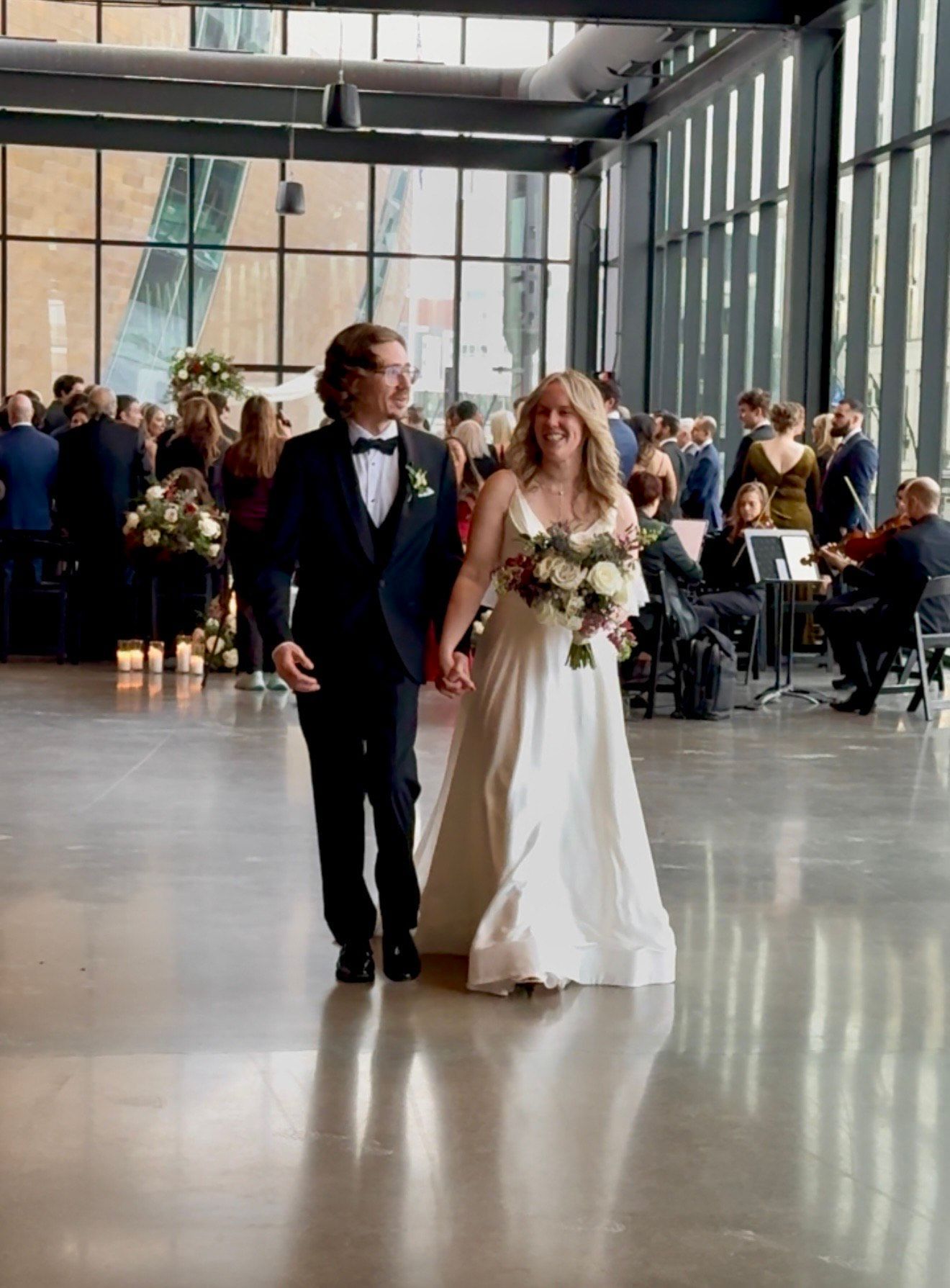Newlyweds walk hand-in-hand at their wedding reception. The bride wears a white dress, the groom a tuxedo. Large windows and guests in the background.