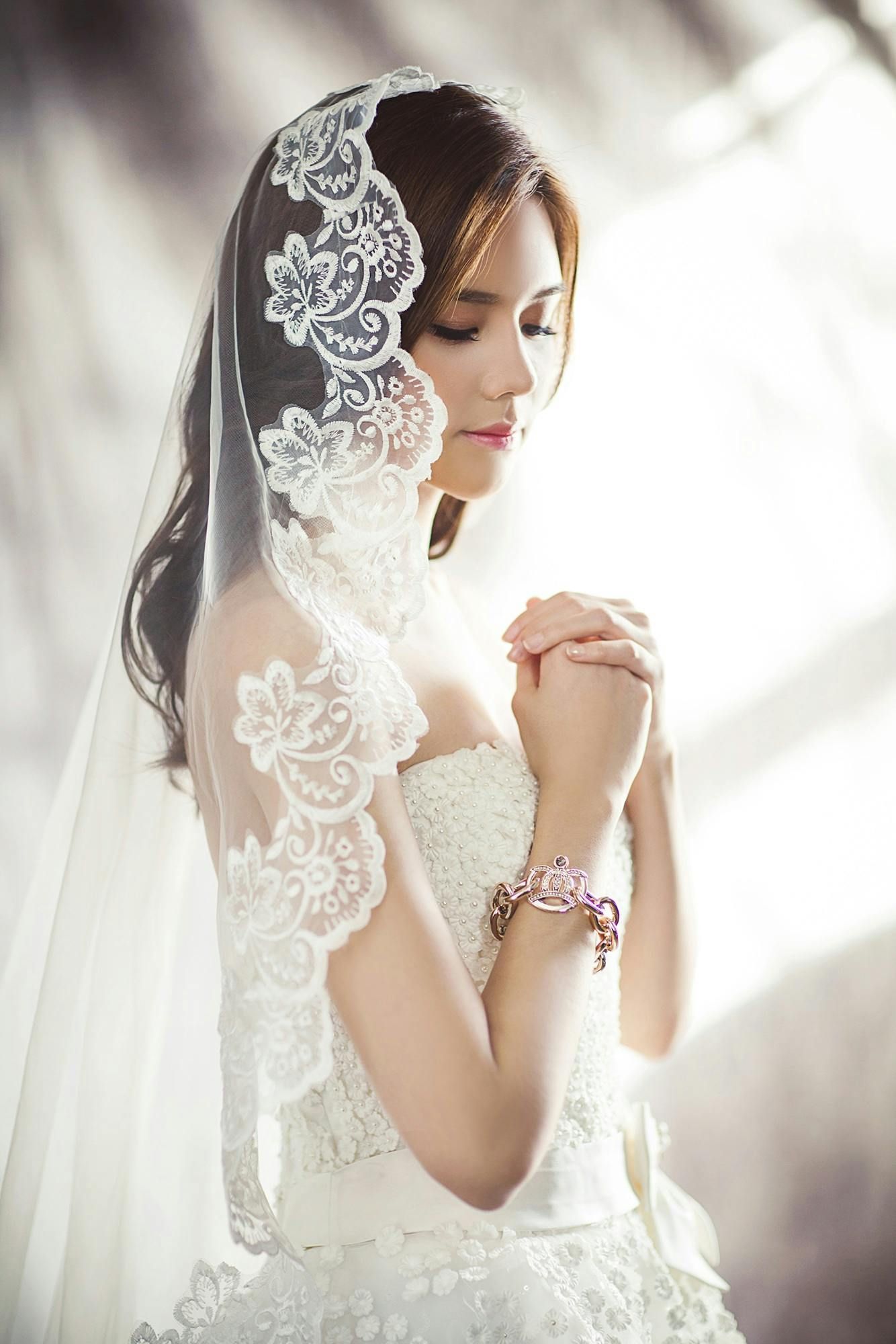 Bride in a white gown and lace veil, with hands clasped in prayer, eyes closed.