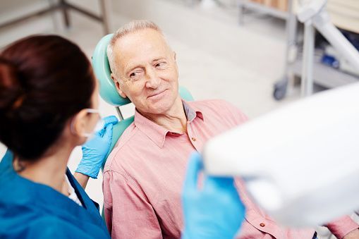 An elderly man is sitting in a dental chair talking to a dentist.