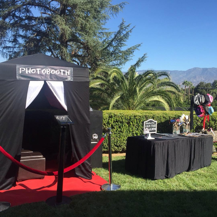 Black photo booth tent with red carpet and velvet ropes beside a decorated table on a sunny lawn