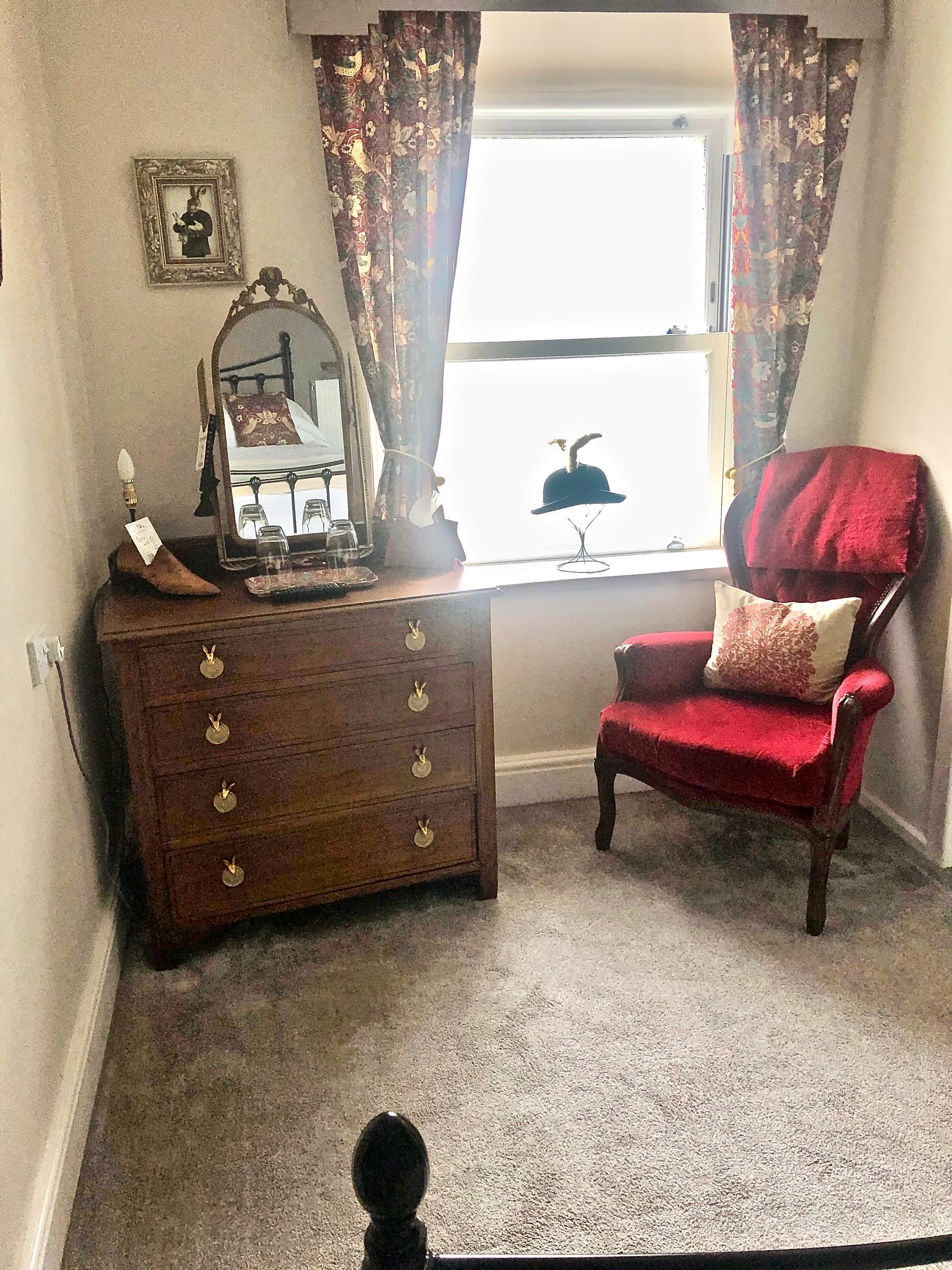 A bedroom with a red chair , dresser , mirror and window.