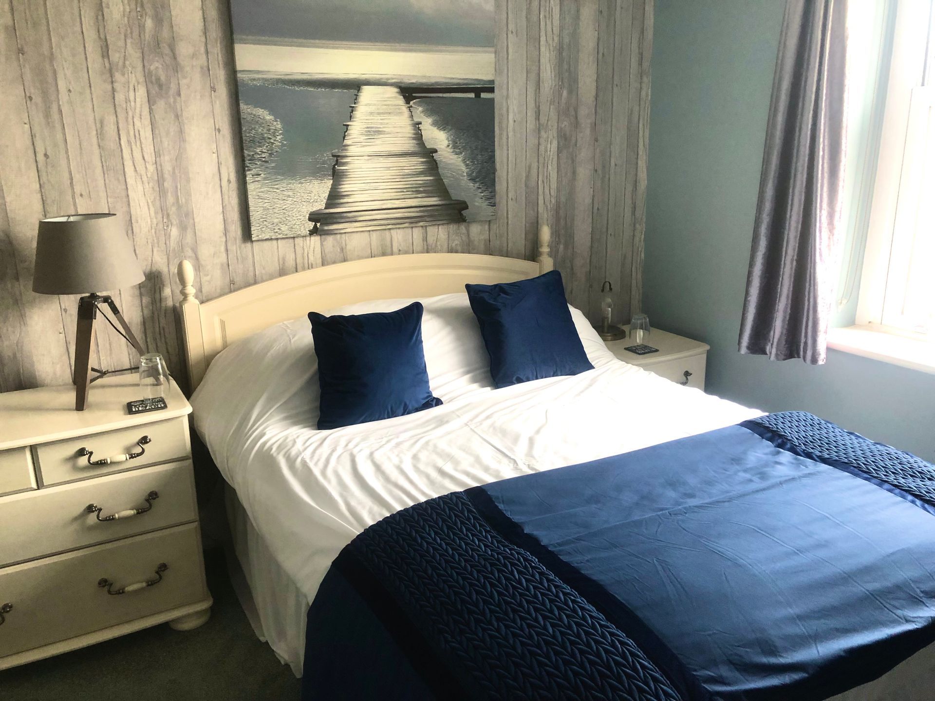 A bedroom with a bed , nightstand , lamp and a painting on the wall.