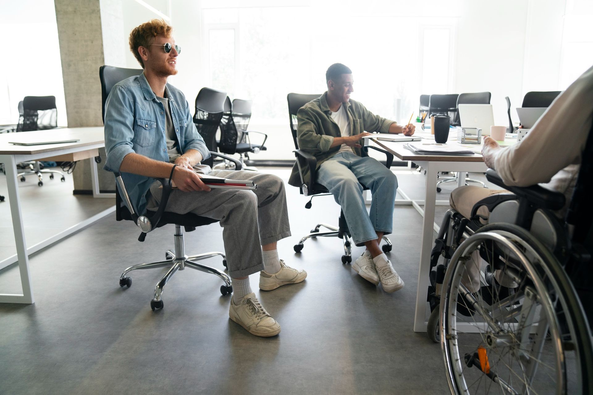 Three people seated at desks in an office; one in a wheelchair, two others are smiling and conversing.