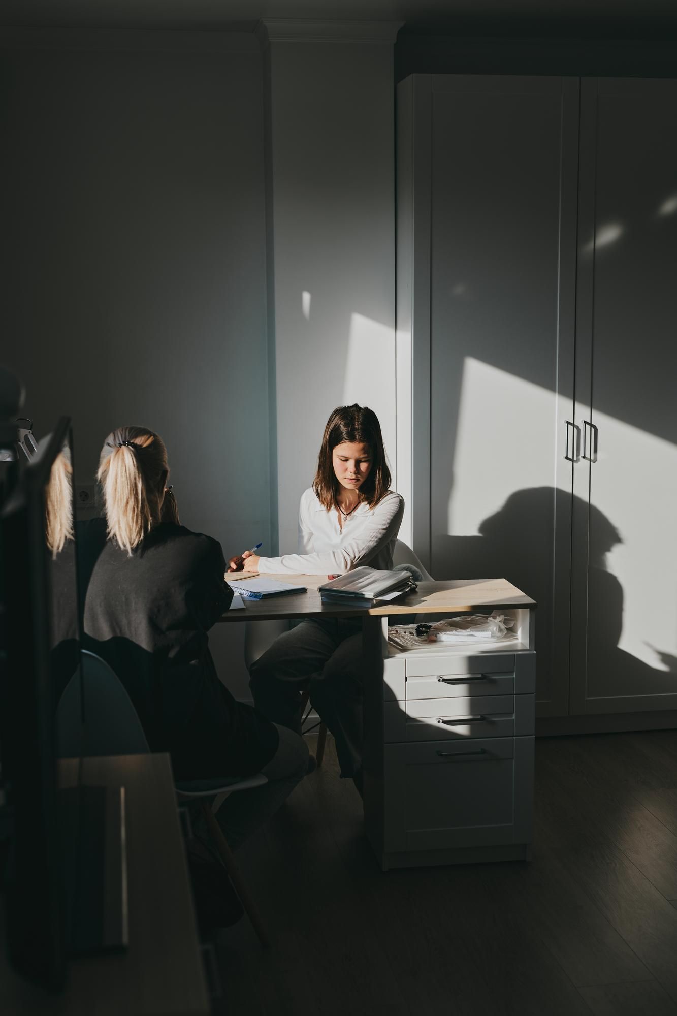Two people sitting at a desk in a sunlit room, one facing the other. One person looks down, the other looks up.