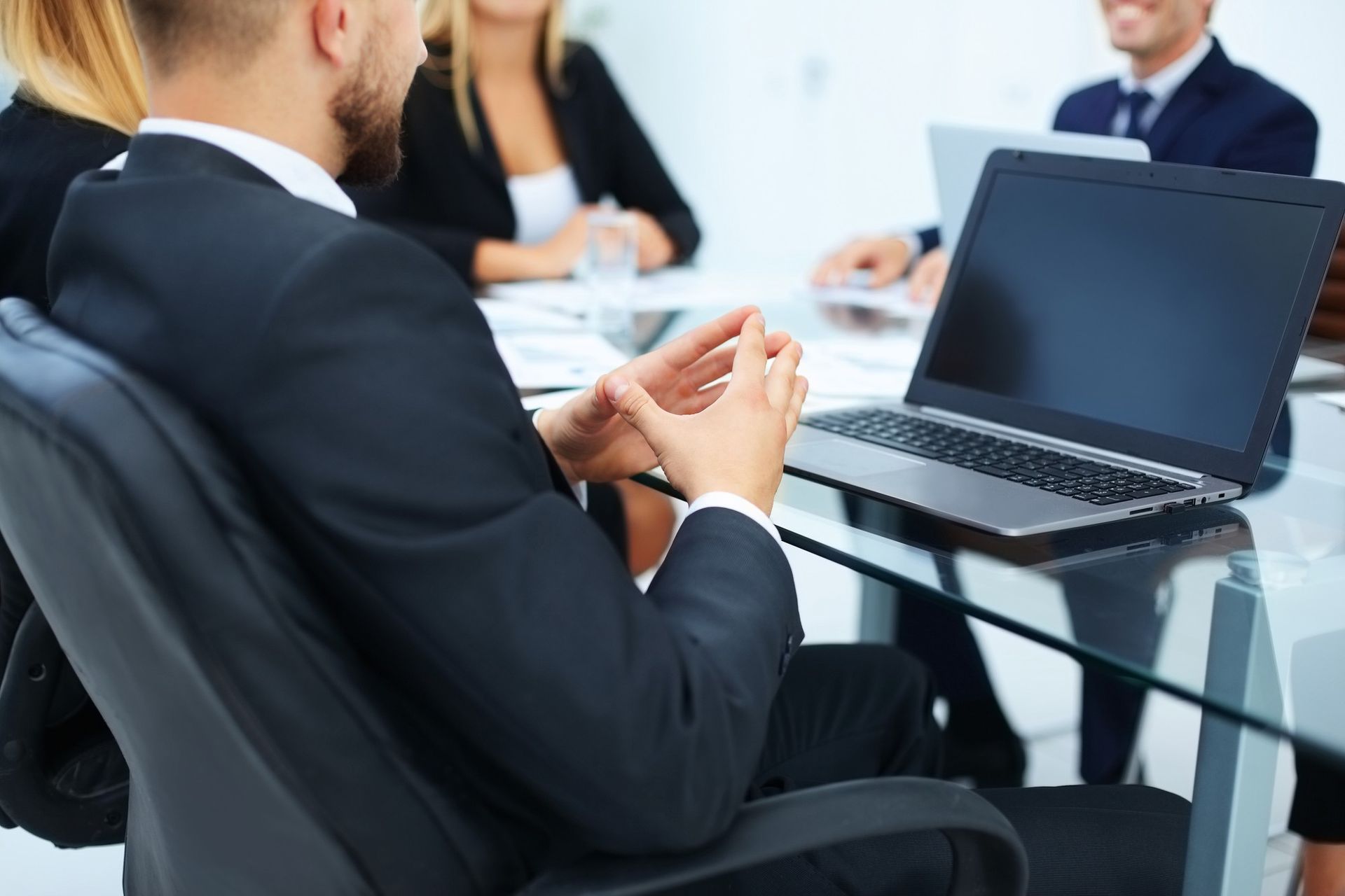 Man in suit at meeting, hands clasped, laptop open on glass table, colleagues blurred in background.