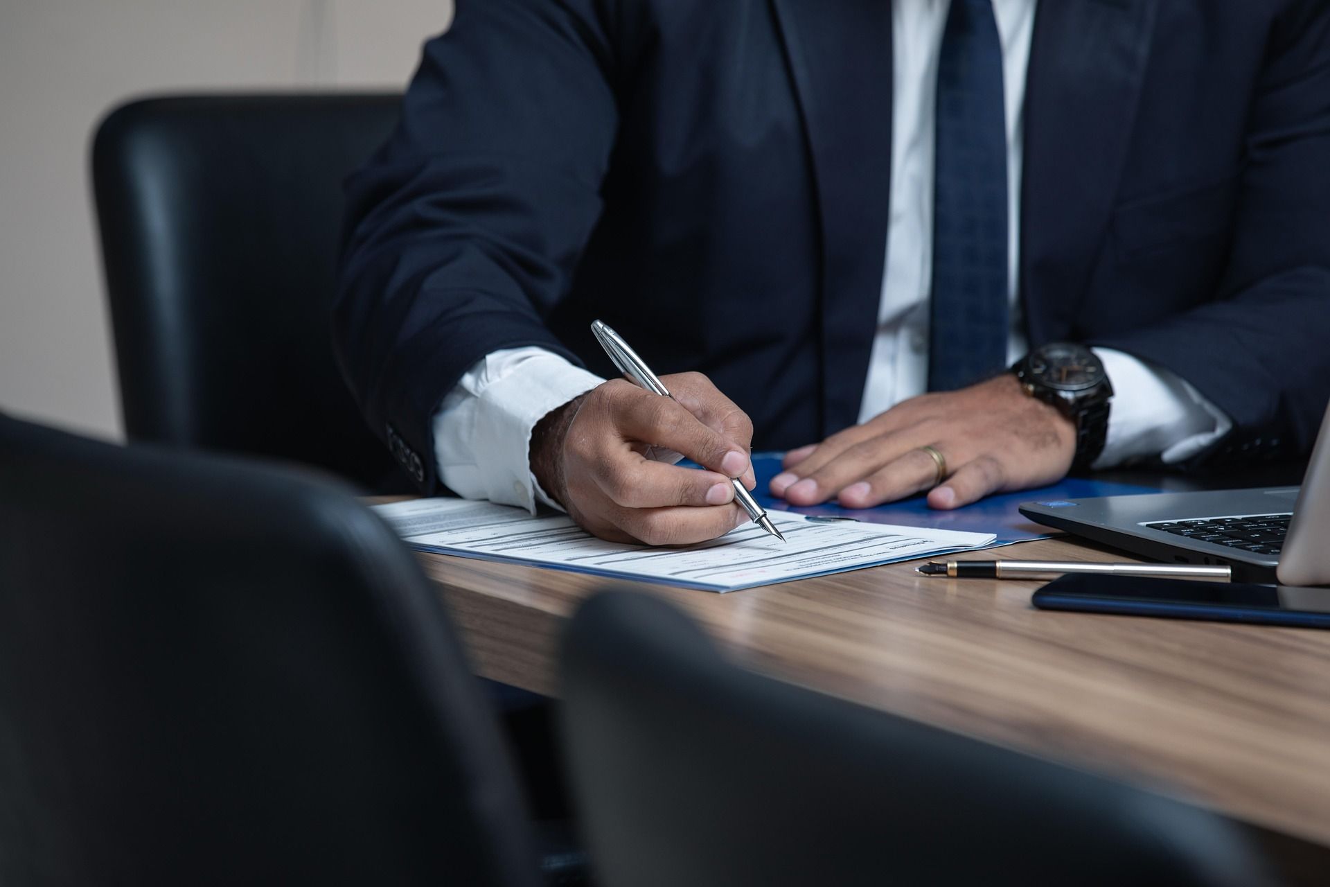Man in suit writing on paper at a desk, laptop nearby.