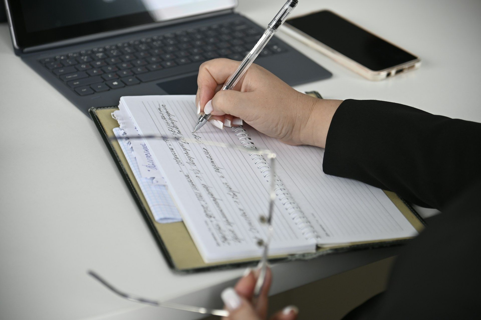 Person writing on a clipboard with a pen, laptop and phone on a white desk.