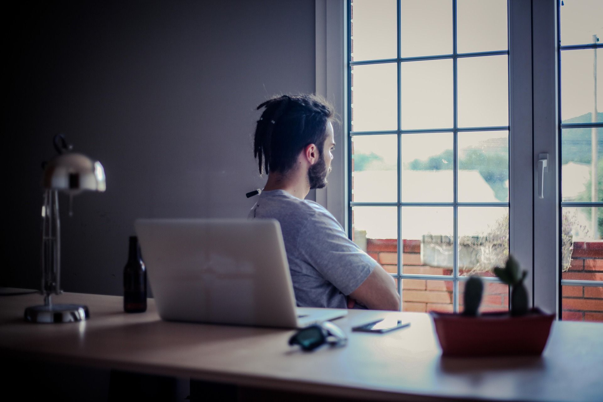 Man with dreadlocks looks out a window from his desk. A laptop, lamp, and cactus sit on the desk.