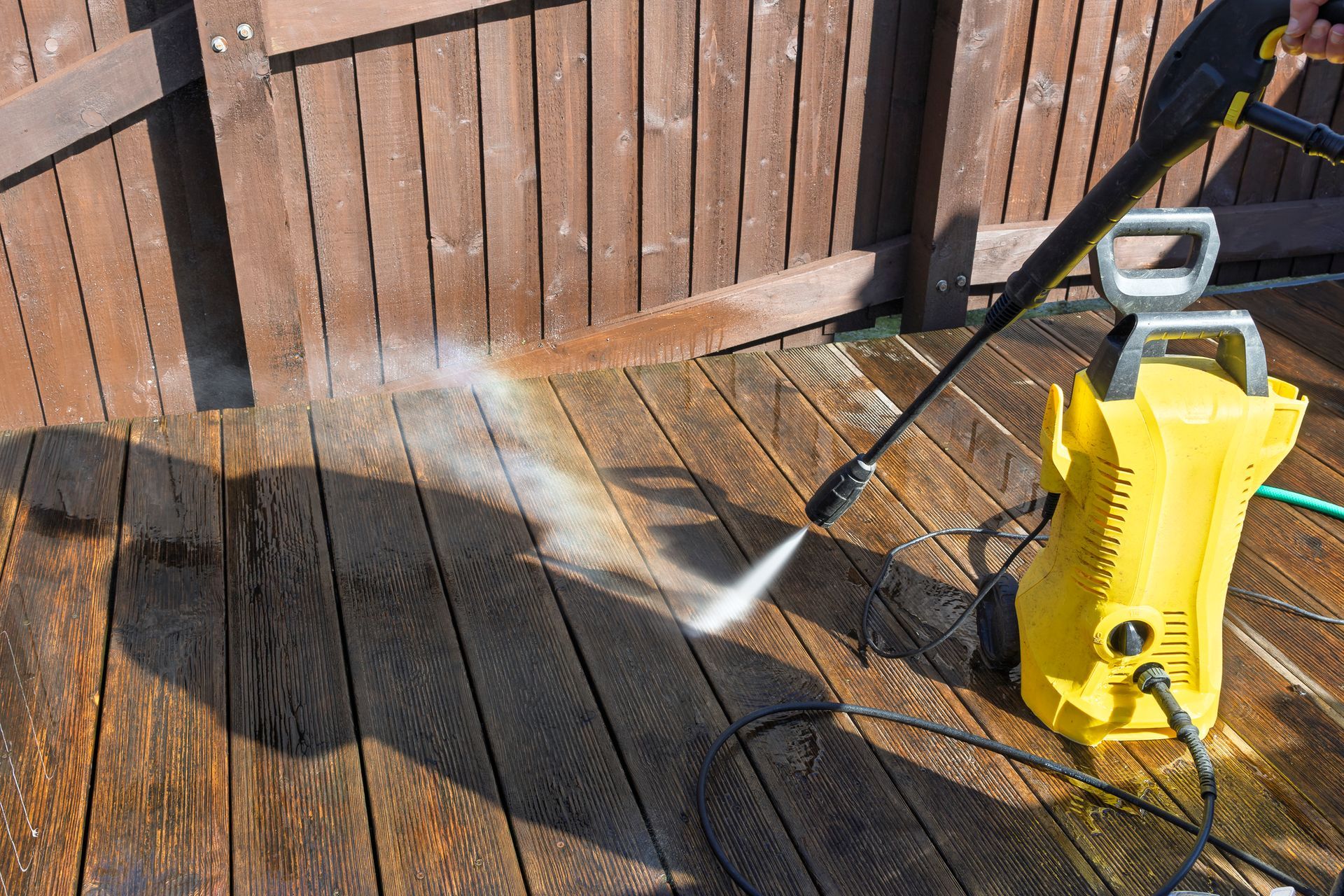 A person pressure washing a wooden deck with a yellow machine, leaving a clean, wet streak.