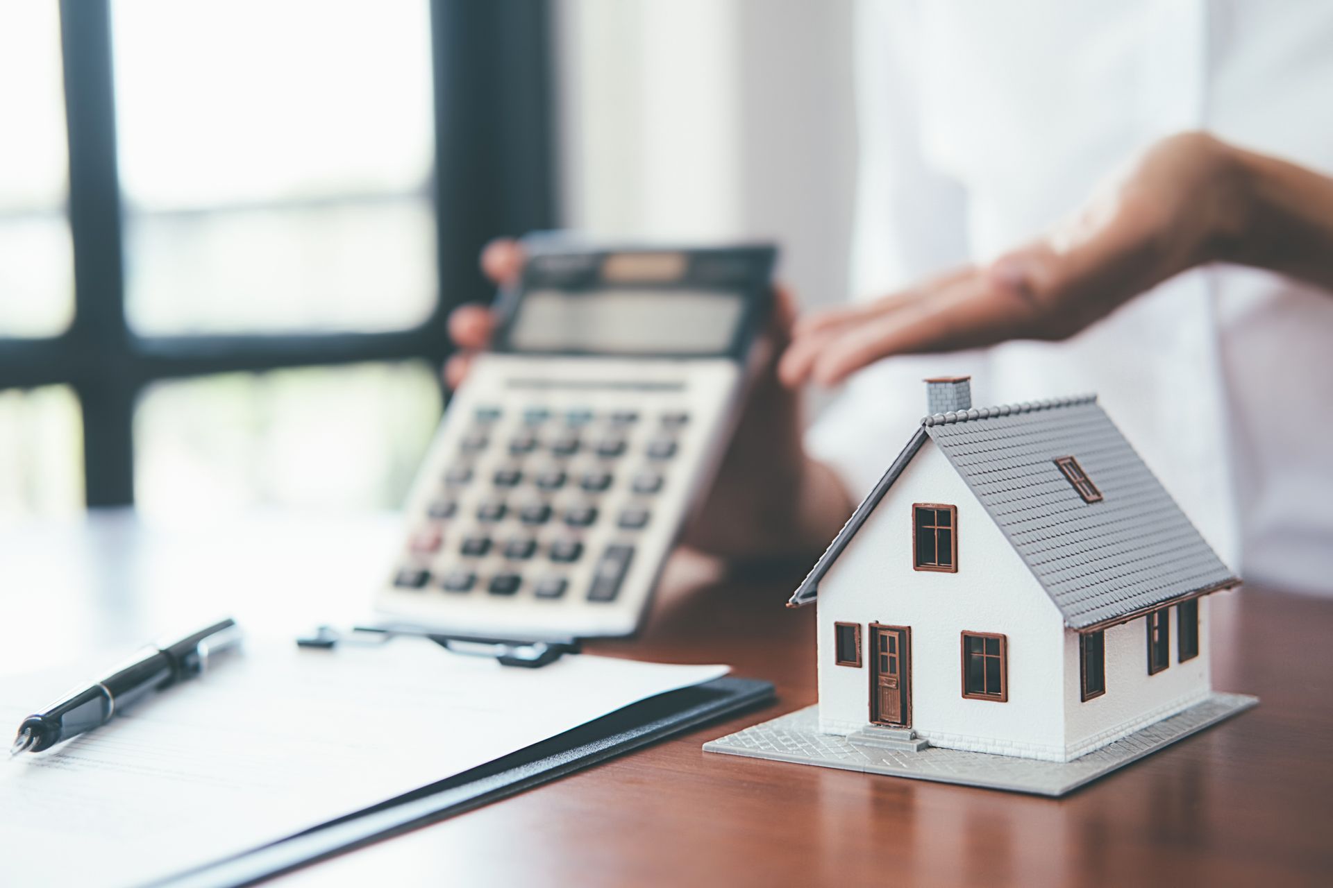 Miniature house, calculator, and document on a table; hand holding calculator, suggesting financial planning.
