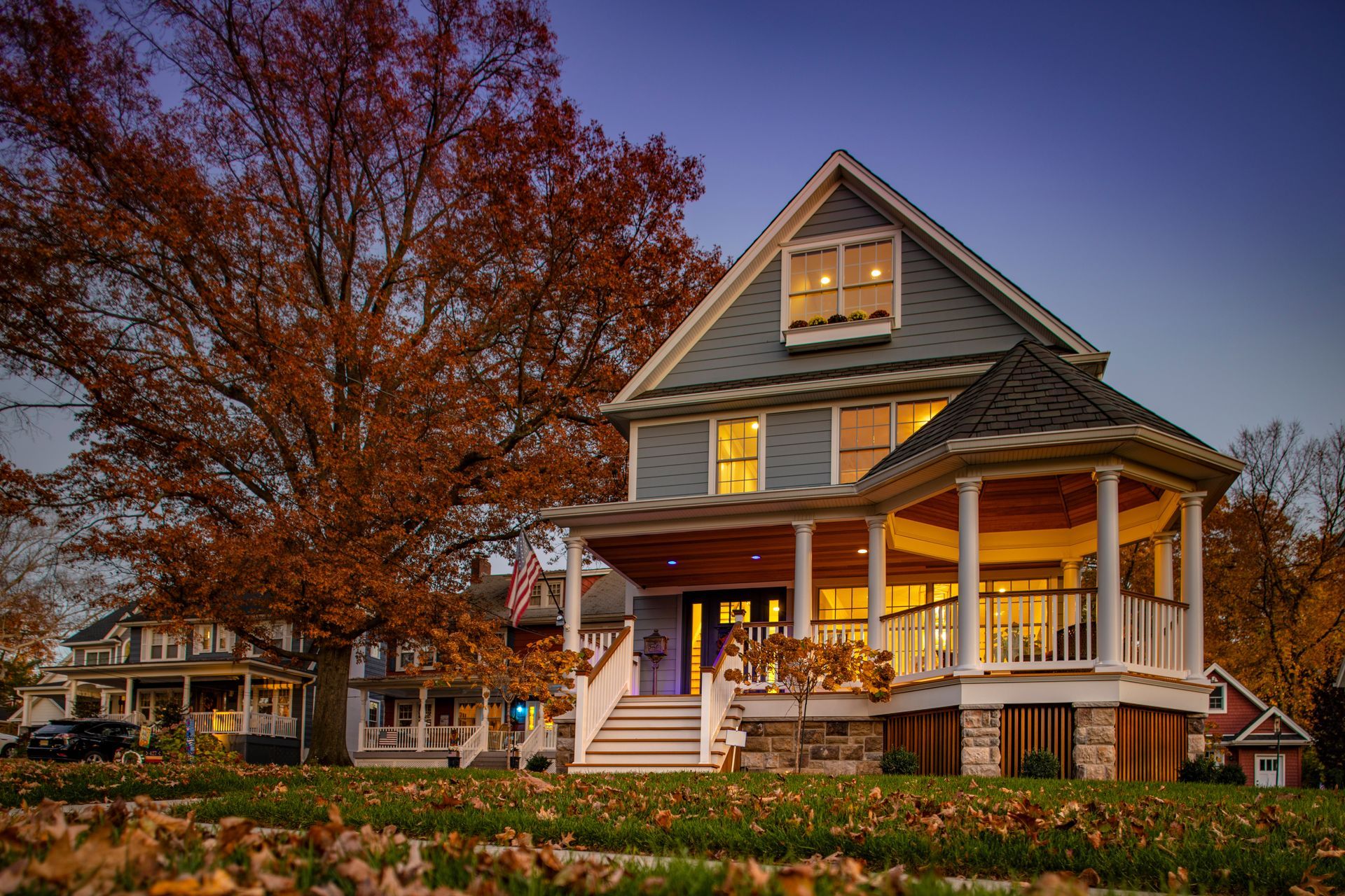 Two-story house with a porch and fall foliage, lit from within, at dusk.