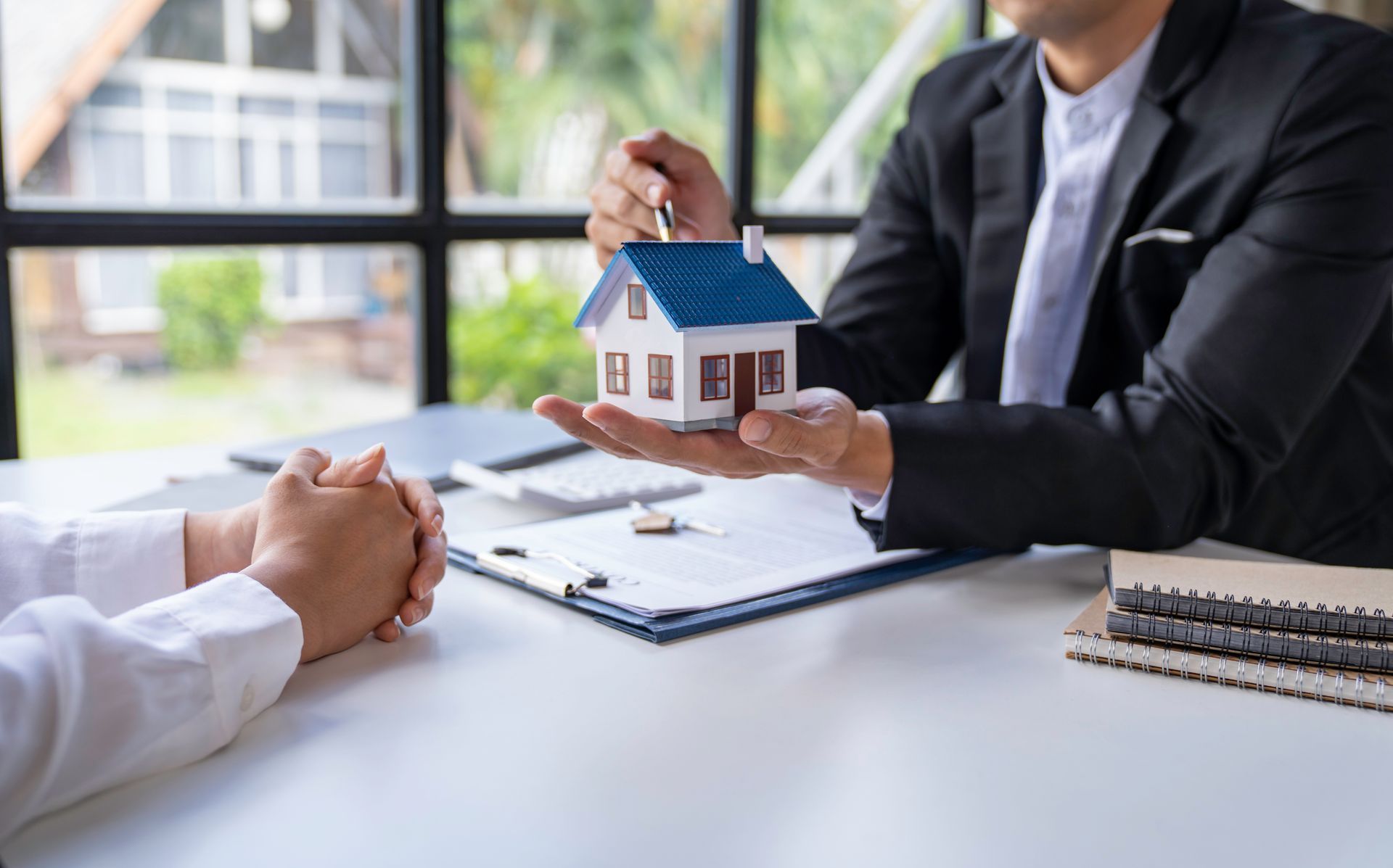 Real estate agent holding house model; client reviews paperwork at a table with a window.