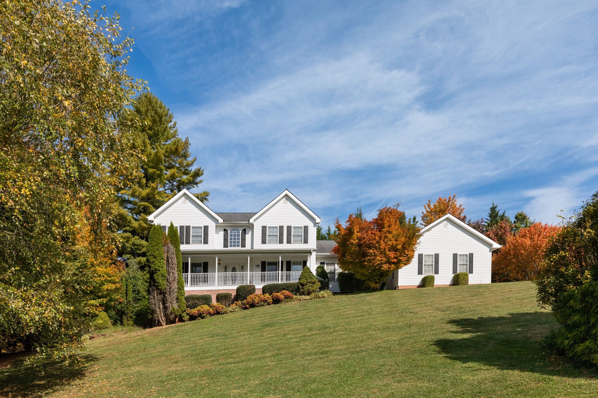 White house with black shutters, on a grassy hill, surrounded by autumn trees, against a blue sky.