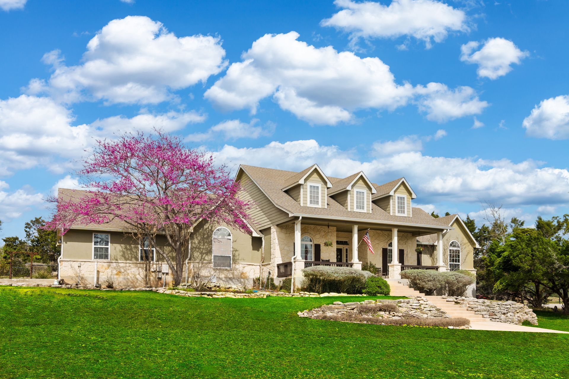 Large beige house with a porch, dormers, and a pink flowering tree on a green lawn under a blue sky with clouds.