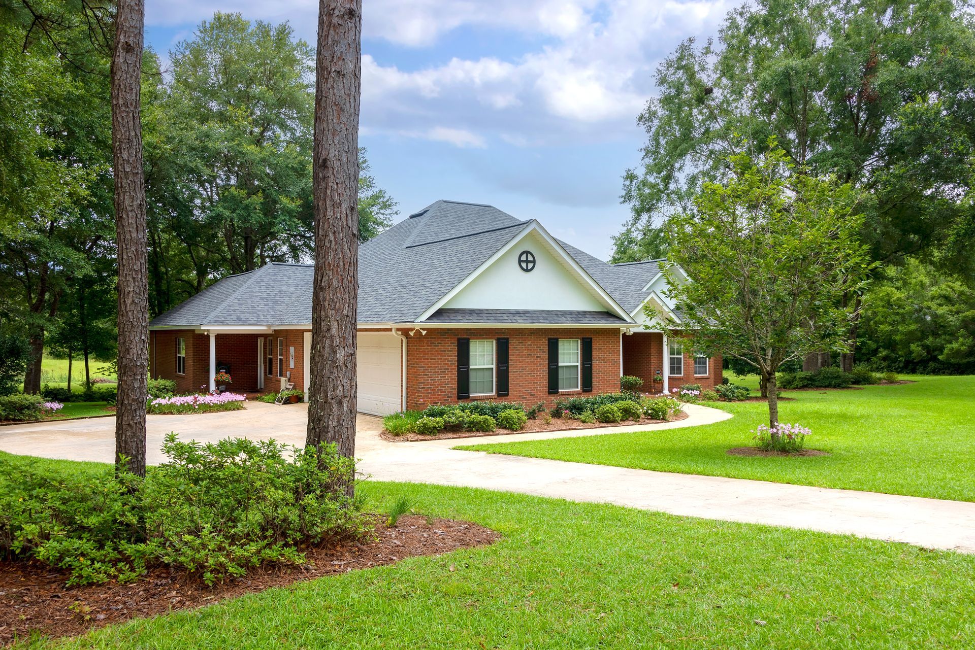 A brick home with a gray roof and circular drive, surrounded by trees and a green lawn under a cloudy sky.