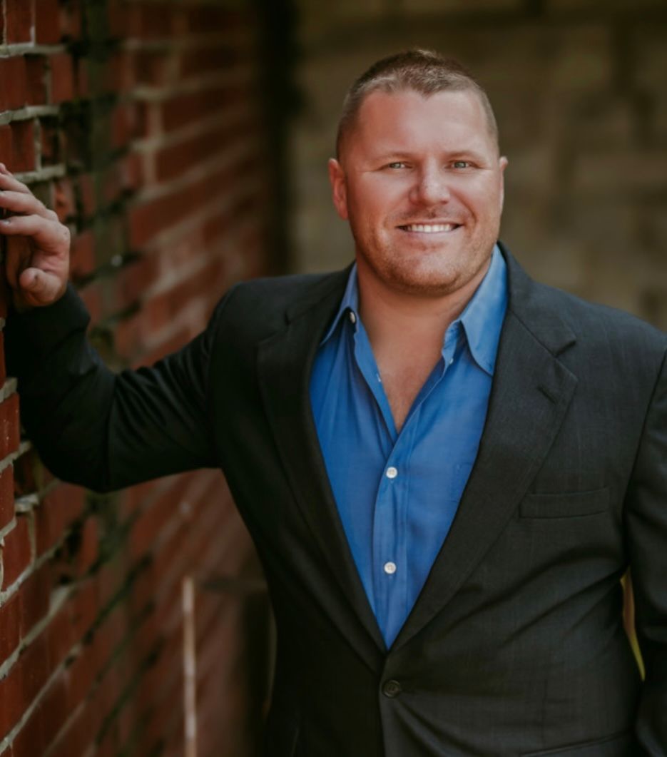 Man in suit and blue shirt smiles, leaning against a brick wall.